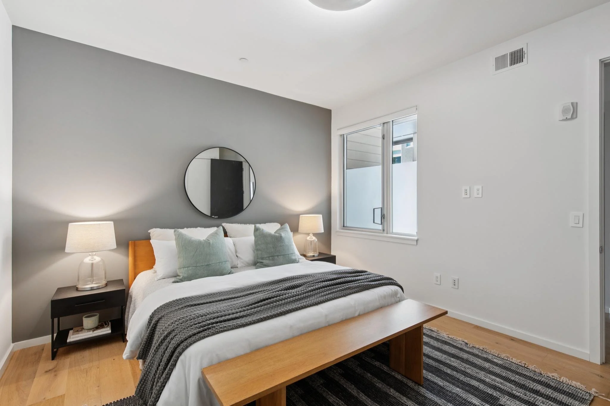 A modern bedroom with a large bed covered in white and gray bedding, accented by two lamps on black bedside tables, a small wooden bench at the foot of the bed, a round mirror on the light gray wall, and a window with open blinds allowing natural lig
