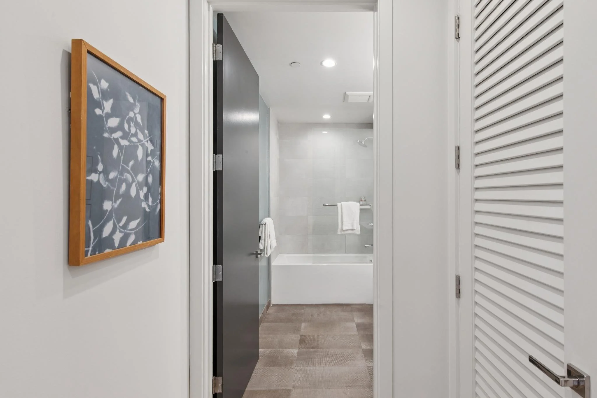 View into a bathroom through an open door, showing a shower with a glass door, white tiled walls, a towel rack, and a white towel. The foreground includes a hallway with a framed picture on the wall and a closed vent.