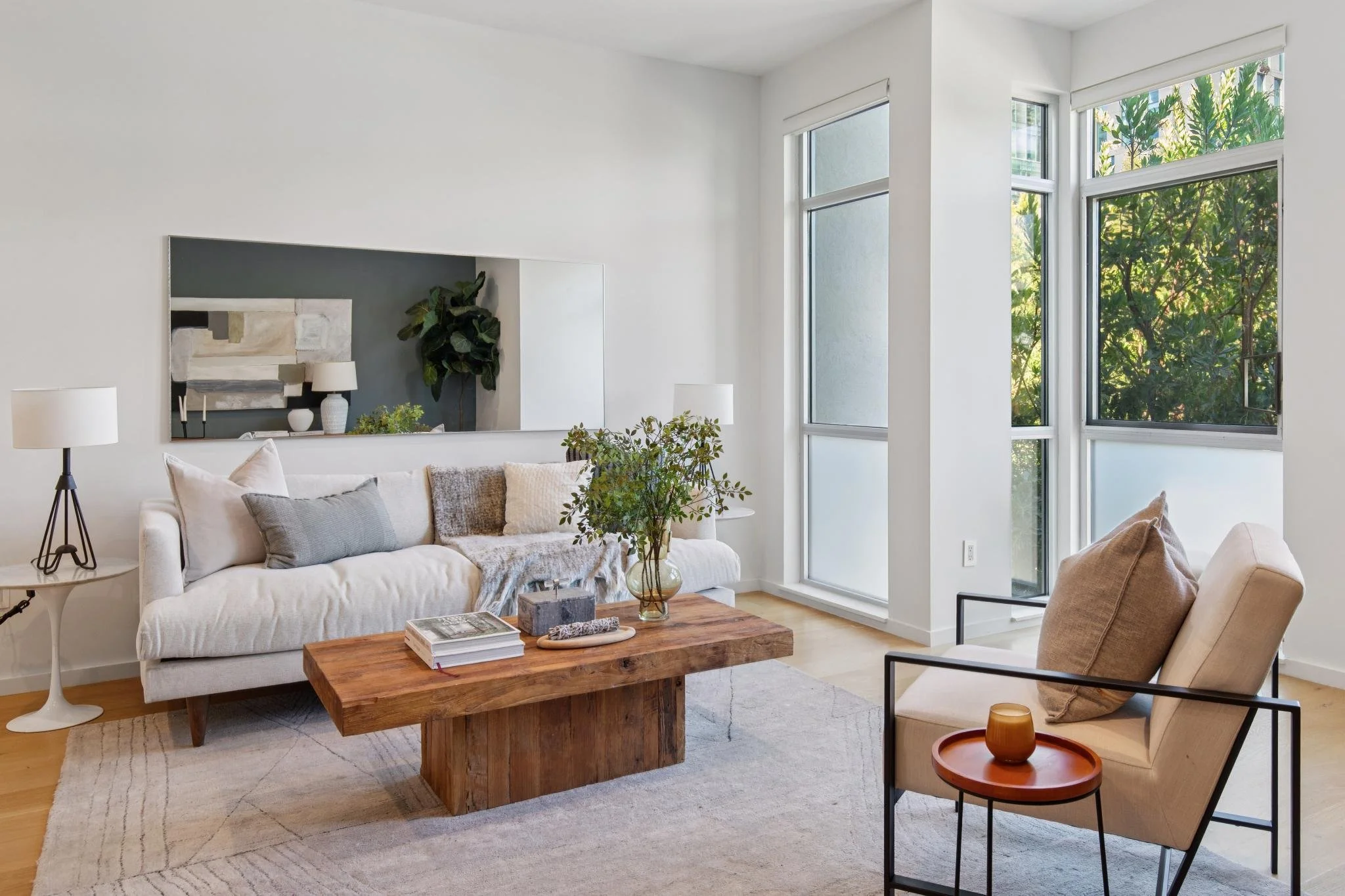 Bright living room with white walls, large windows with greenery outside, a white couch with pillows, a wooden coffee table with books and decor, a side table with a lamp, and a beige armchair with a pillow.
