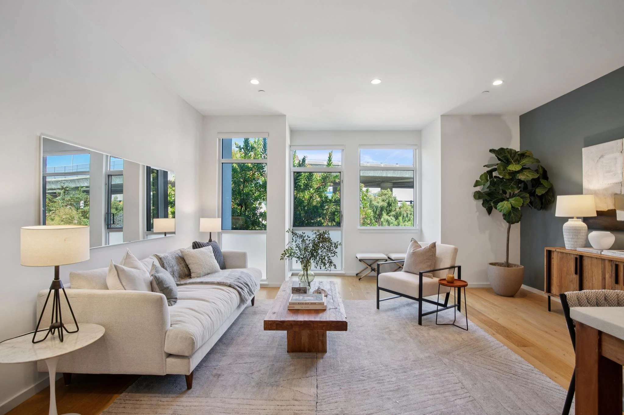 A modern living room with white walls and large windows showing greenery outside. It features a white sofa with pillows, a wooden coffee table, a side table with a lamp, and a tall potted plant. The room has a neutral color palette with cozy and styl