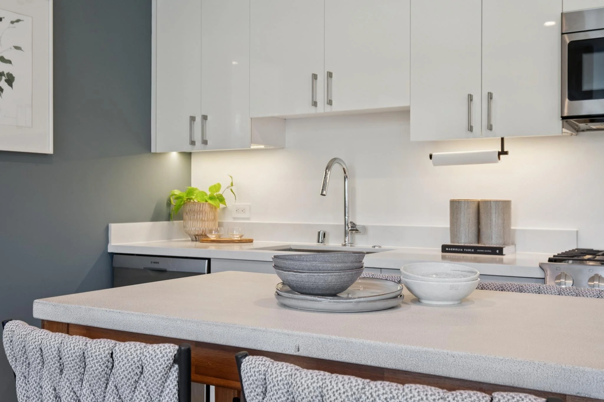 A modern kitchen with white cabinets, a white countertop, a silver faucet, and a potted green plant. There are bowls and a tray on the counter, and a book titled "Magnolia Table" is visible. The backsplash is light-colored, and the overall style is m