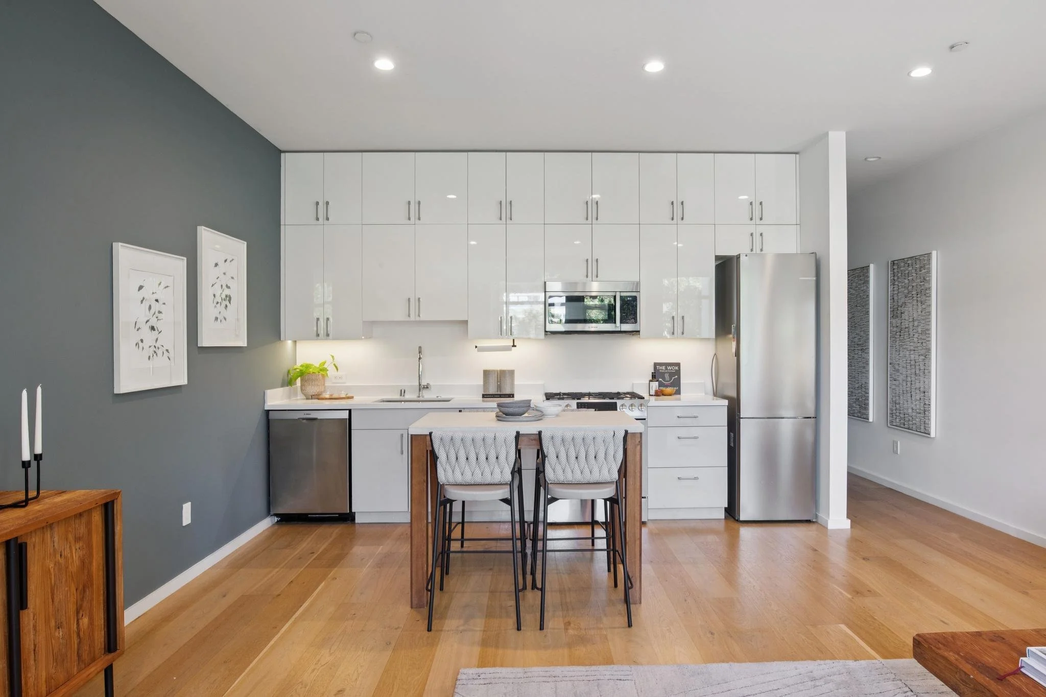Modern kitchen with white cabinets, stainless steel appliances, and a small island with two chairs. Hardwood floors, decorative wall art, and a plant on the counter.