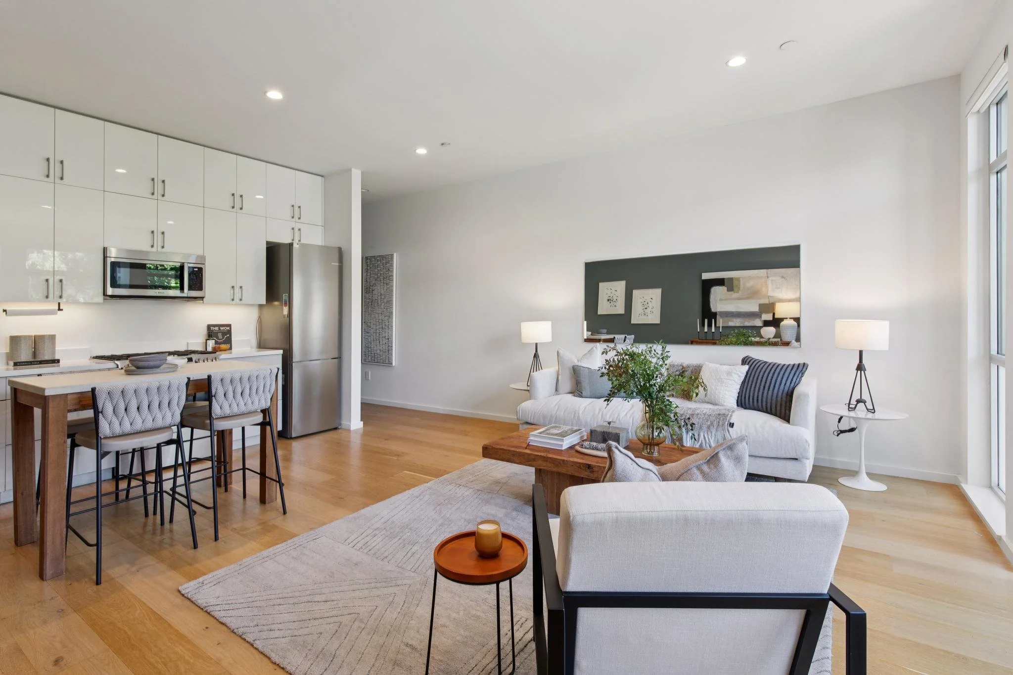 Open-concept living room and kitchen with white walls, hardwood floors, and large windows; living area features white sofa with black and white striped pillows, two side tables with lamps, a wooden coffee table with books and a plant; kitchen has whi