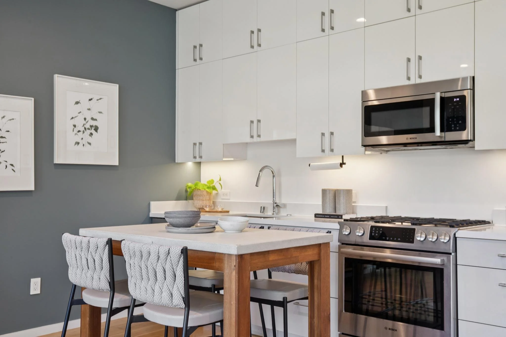 Modern kitchen with white cabinets, stainless steel microwave, gas stove, white countertop, gray accent wall with framed botanical prints, dining table with gray and black chairs, and green potted plant, in a well-lit space.
