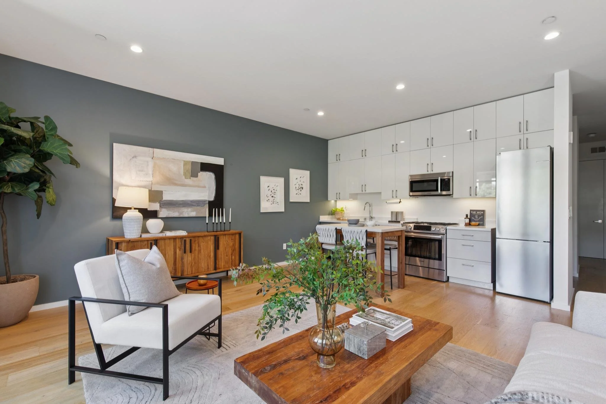 Open-concept living room and kitchen with modern decor, including a white armchair, a wooden coffee table with plants, a grey accent wall, and white kitchen cabinets with stainless steel appliances.