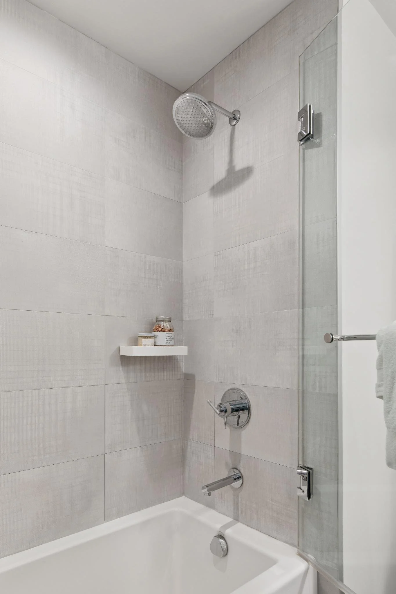 A clean, modern bathroom shower area with a white bathtub, a glass shower door, a round shower head, and a soap shelf with bottles of soap or shampoo.