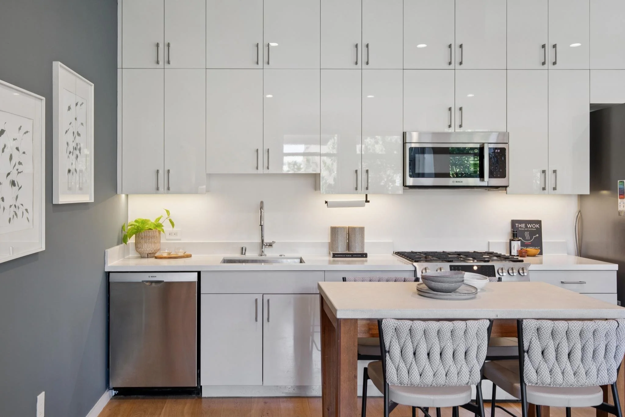 Modern kitchen with white cabinets, stainless steel microwave, and a white countertop. There's a potted plant on the left, a paper towel holder, and a stove with a cooking book on the right. A dining table with chairs is in the foreground.