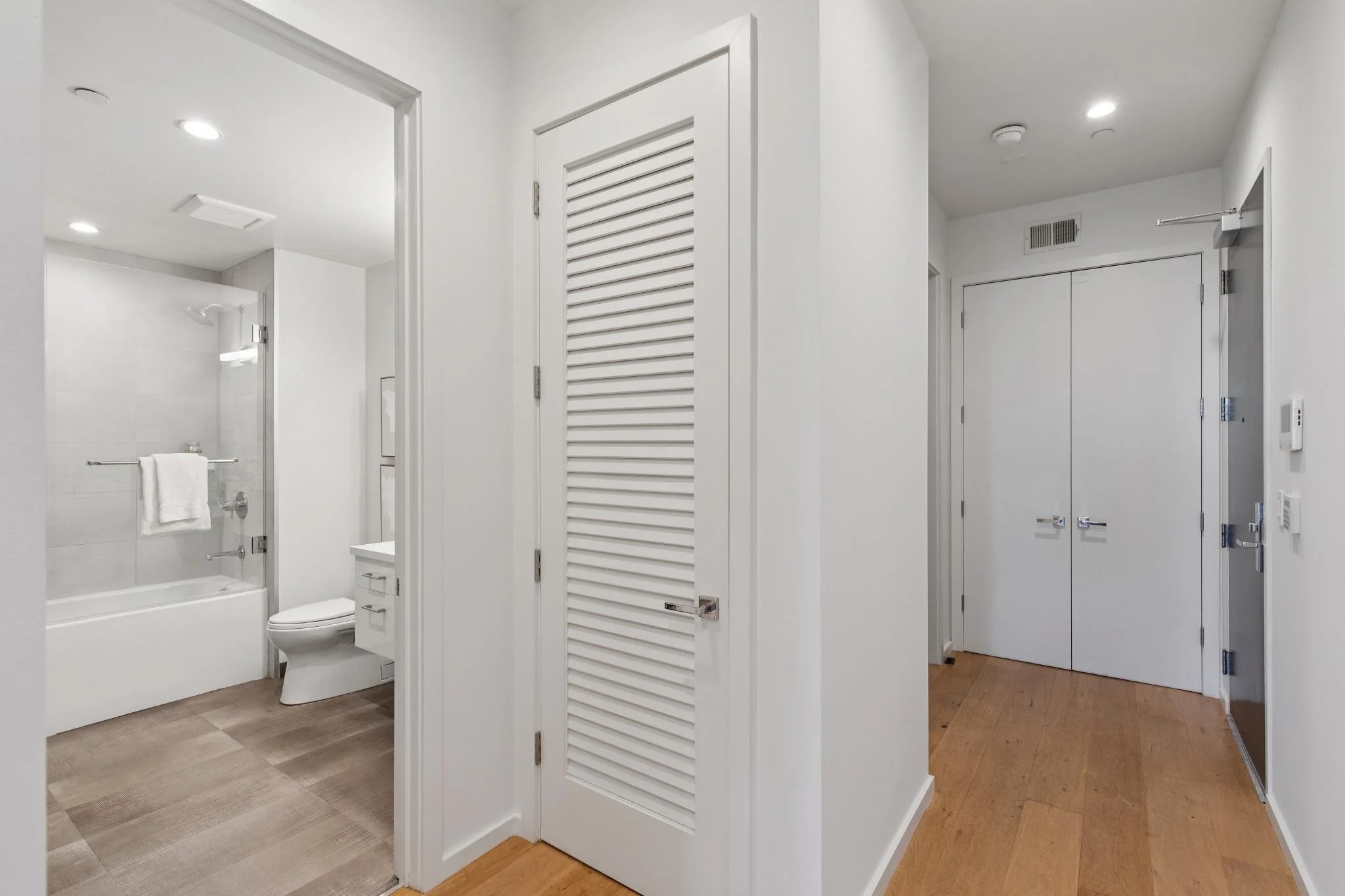 Modern bathroom with white walls, a glass-enclosed shower, a toilet, and a white cabinet, with wooden flooring visible in the hallway.
