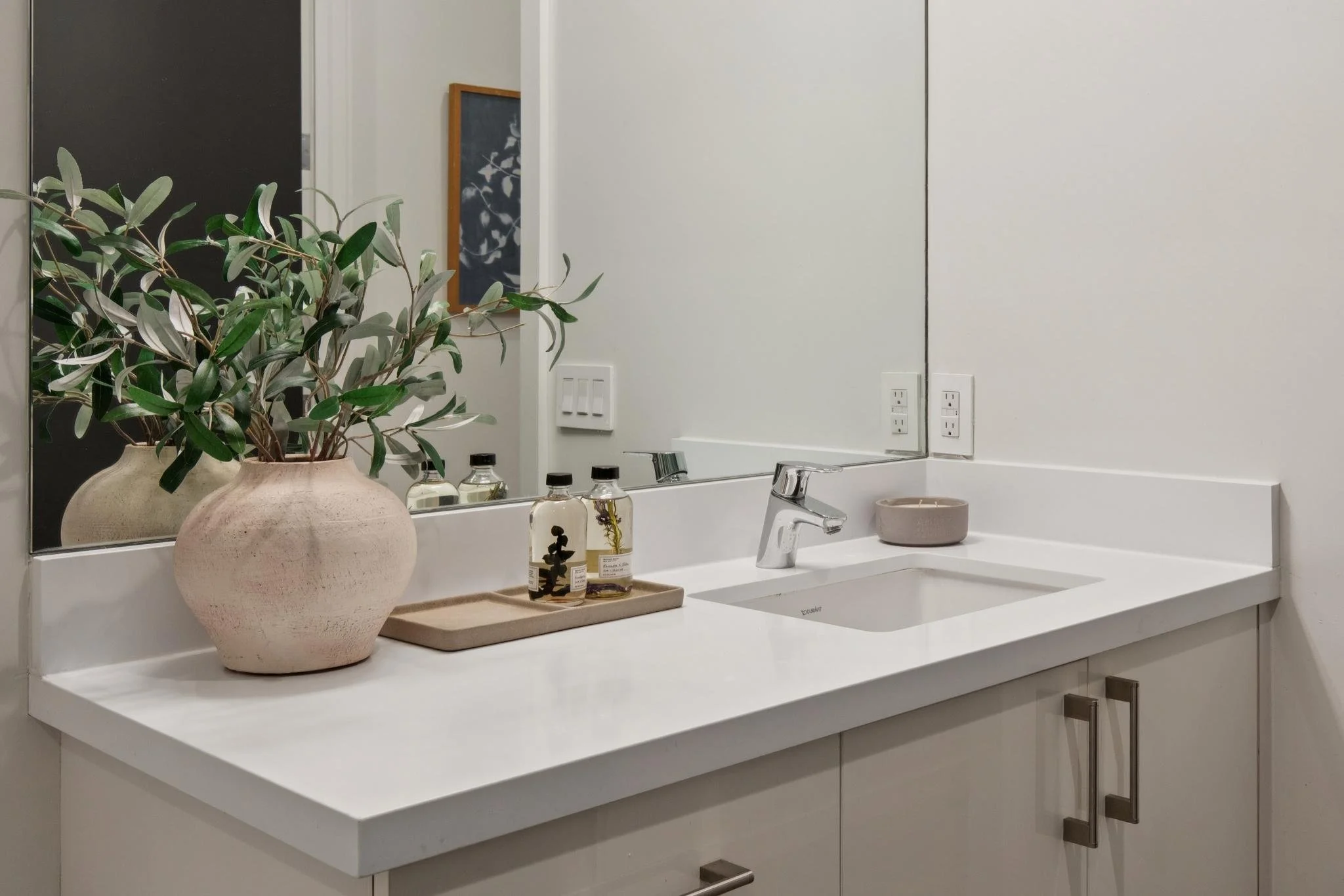 A clean white bathroom vanity with a large mirror, a small tray holding two skincare bottles, a beige soap dish, and a potted plant with green and silver leaves. The background includes a blackboard or note board on the wall and multiple electrical o