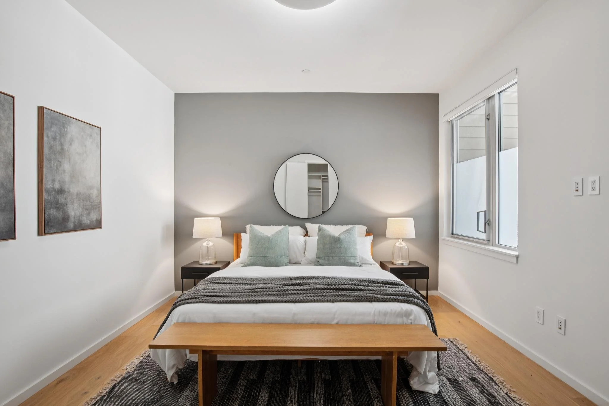 Modern bedroom with white walls, a bed with white and mint green pillows, black nightstands, two table lamps, artwork on the wall, and a large window.
