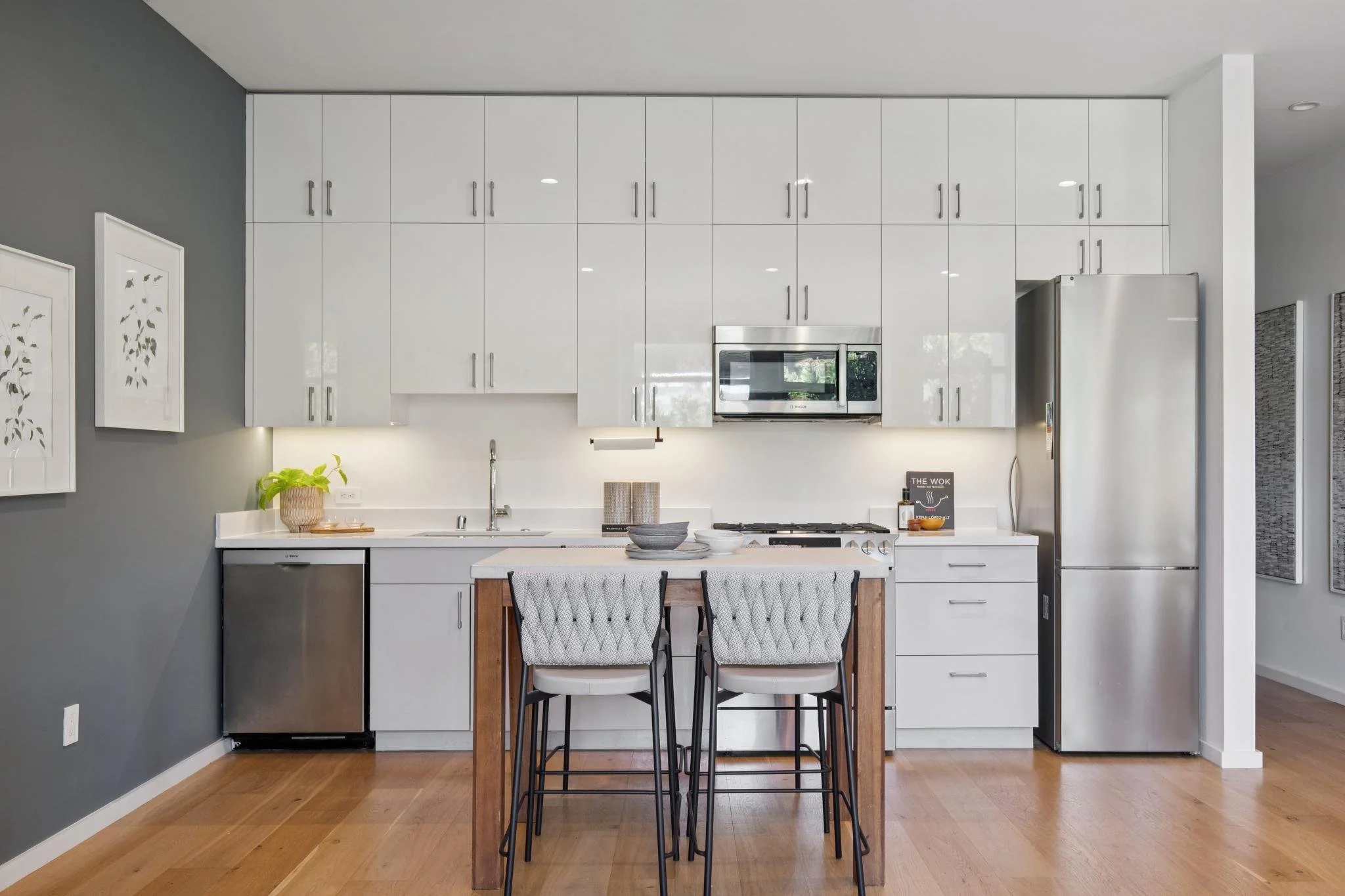 Modern kitchen with white cabinets, stainless steel appliances, and a wooden kitchen island with four black metal chairs, hardwood flooring, and decorative wall art.