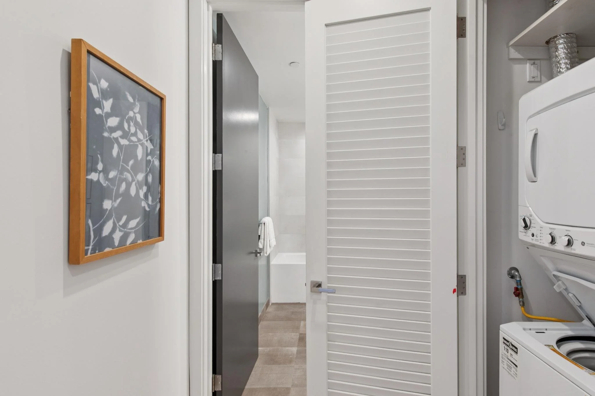 A laundry room with a washing machine on the right, a white louvered door, a hanging towel, a framed picture of leaves on the wall, and a door leading to another room.