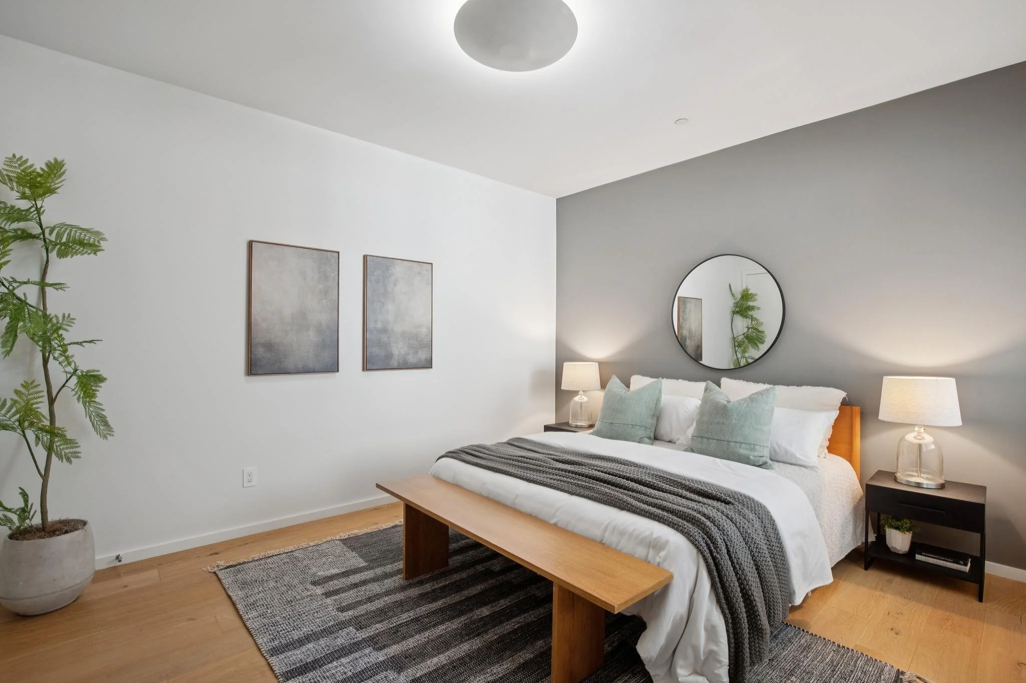 Modern bedroom with white and gray walls, a bed with white linens and light green pillows, wooden bench at foot of bed, black nightstands with lamps, large mirror, framed abstract art, and potted plant in corner.