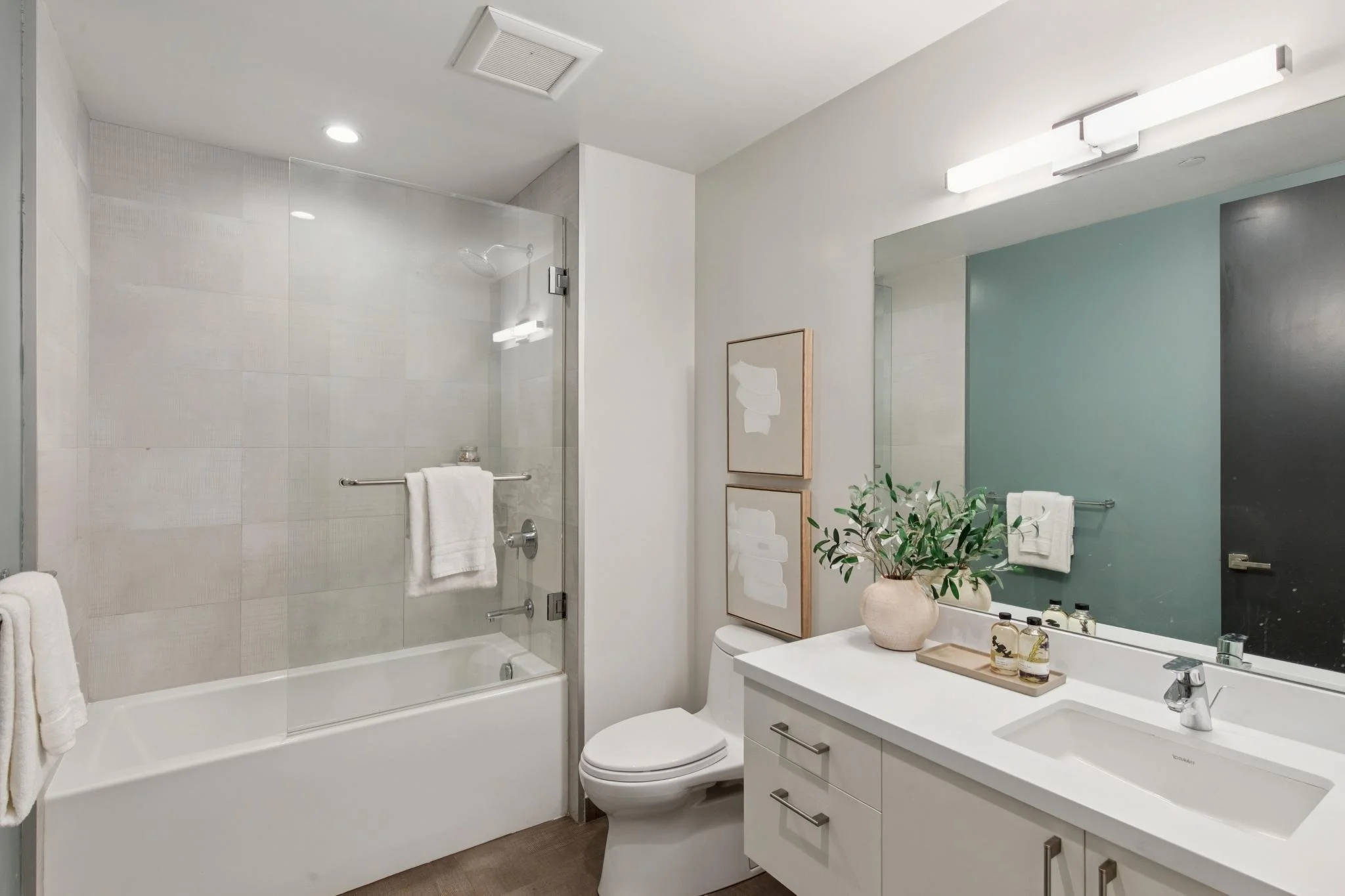 Modern bathroom featuring a glass-enclosed shower with white tiles, a white vanity with a sink, a toilet, a large mirror, and a potted plant on the counter. There are framed artwork and towel racks.