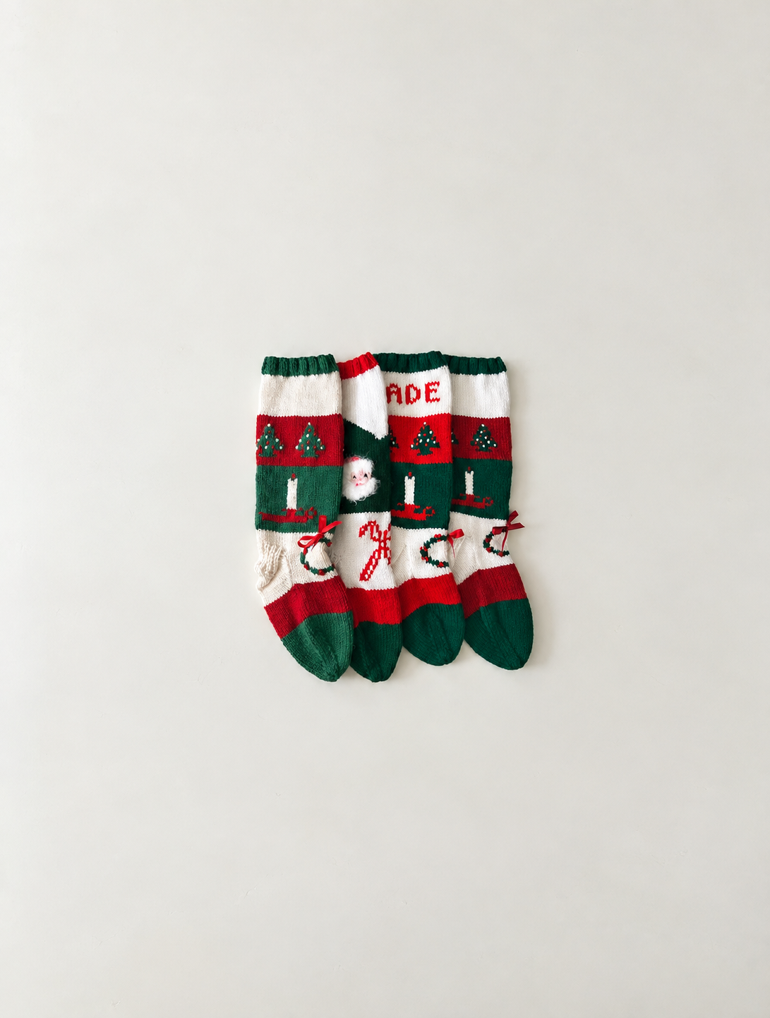 Three Christmas-themed socks with red, green, and white patterns, including Christmas trees, a Santa face, and the word 'MADE' on one sock, tied with small red bows, laid out on a plain white surface.