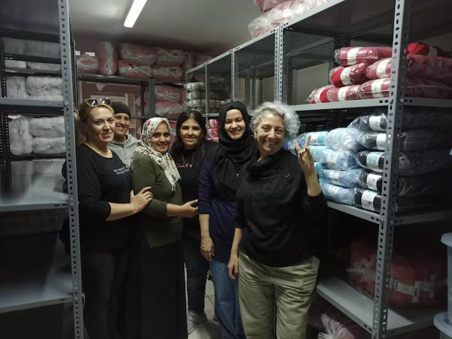 Six women standing in a storage room with shelves filled with blankets and bedding, smiling and posing for the photo.