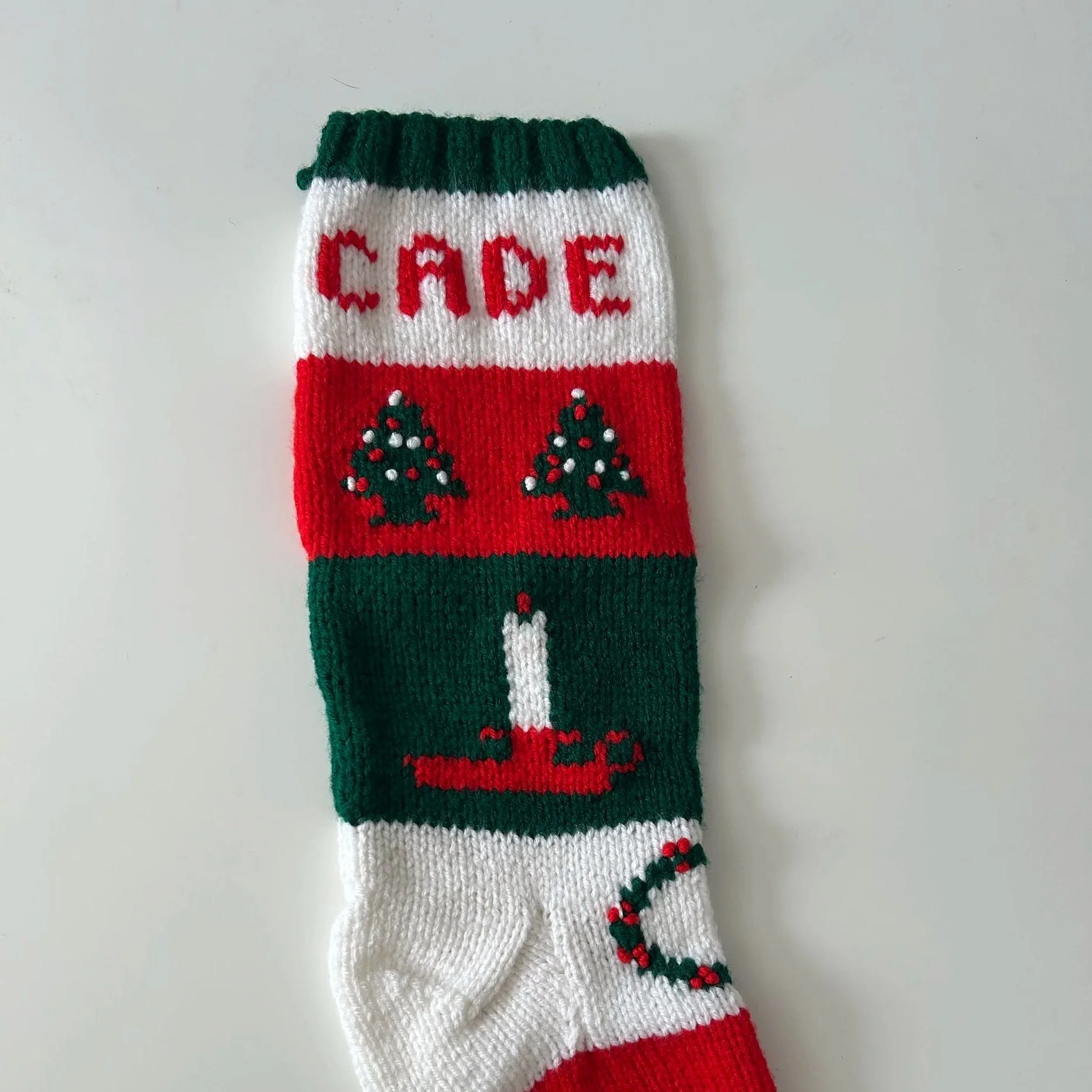 A Christmas stocking with the word 'CACE' embroidered at the top, decorated with Christmas trees and holly berries, in red, green, white, and red colors.