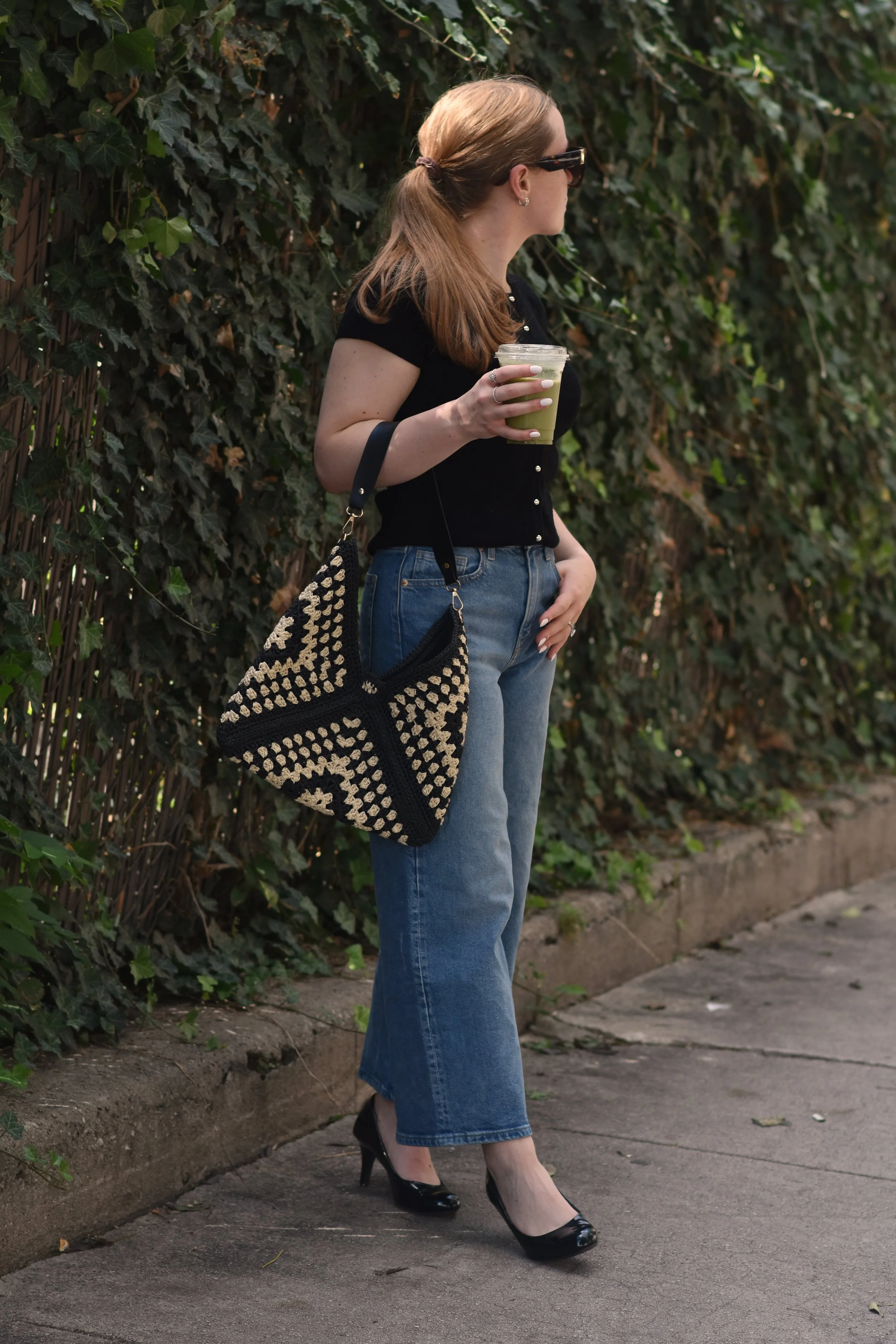 A woman standing on a sidewalk near a hedge, holding a green smoothie in a clear cup. She is wearing black high heels, a black top with short sleeves, blue jeans, and sunglasses. Her light brown hair is styled in a ponytail, and she has a patterned bag hanging from her shoulder.