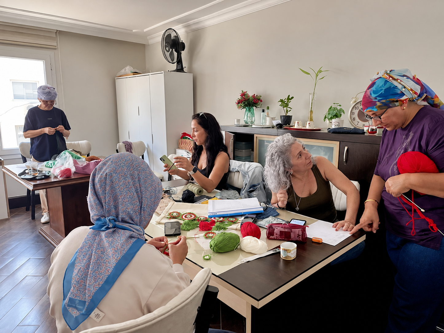 Women gathered in a kitchen or craft room knitting, sewing, and talking, with yarn, knitting supplies, and a laptop on the table.