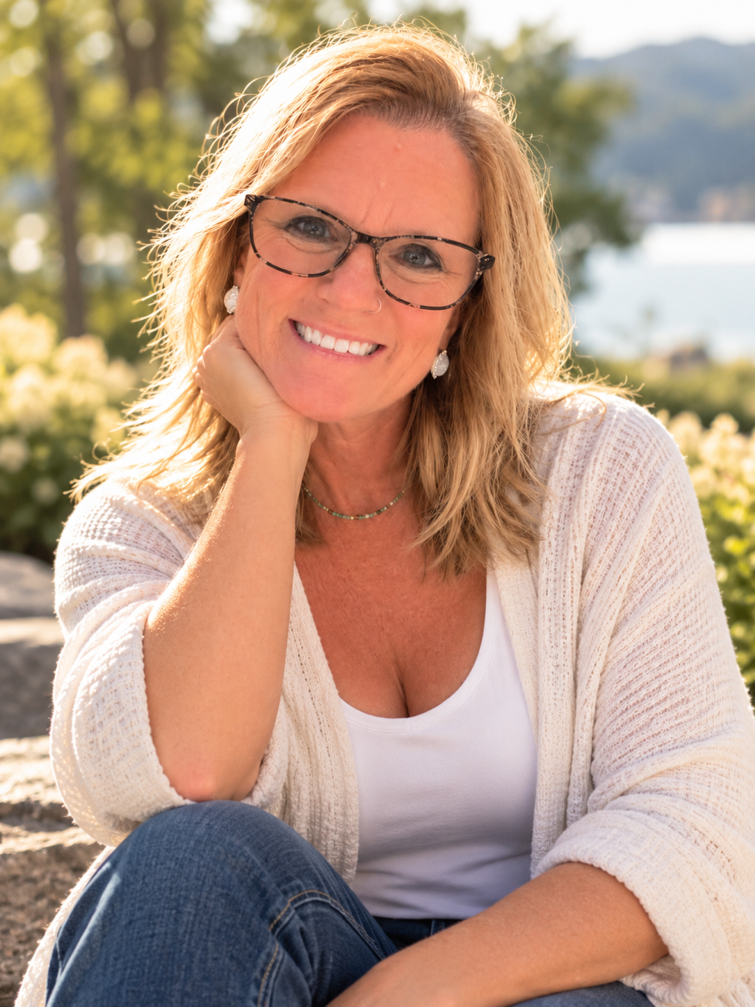 A smiling woman with blonde hair wearing glasses, earrings, and a white top, sitting outdoors with trees and water in the background.