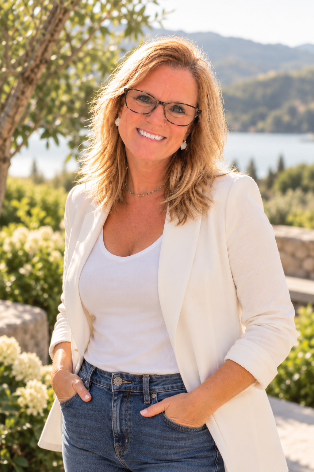 A woman with blonde, wavy hair and glasses standing outdoors near water, smiling. She is wearing a white blazer, a white top, and jeans, with a background of trees, bushes, and mountains.