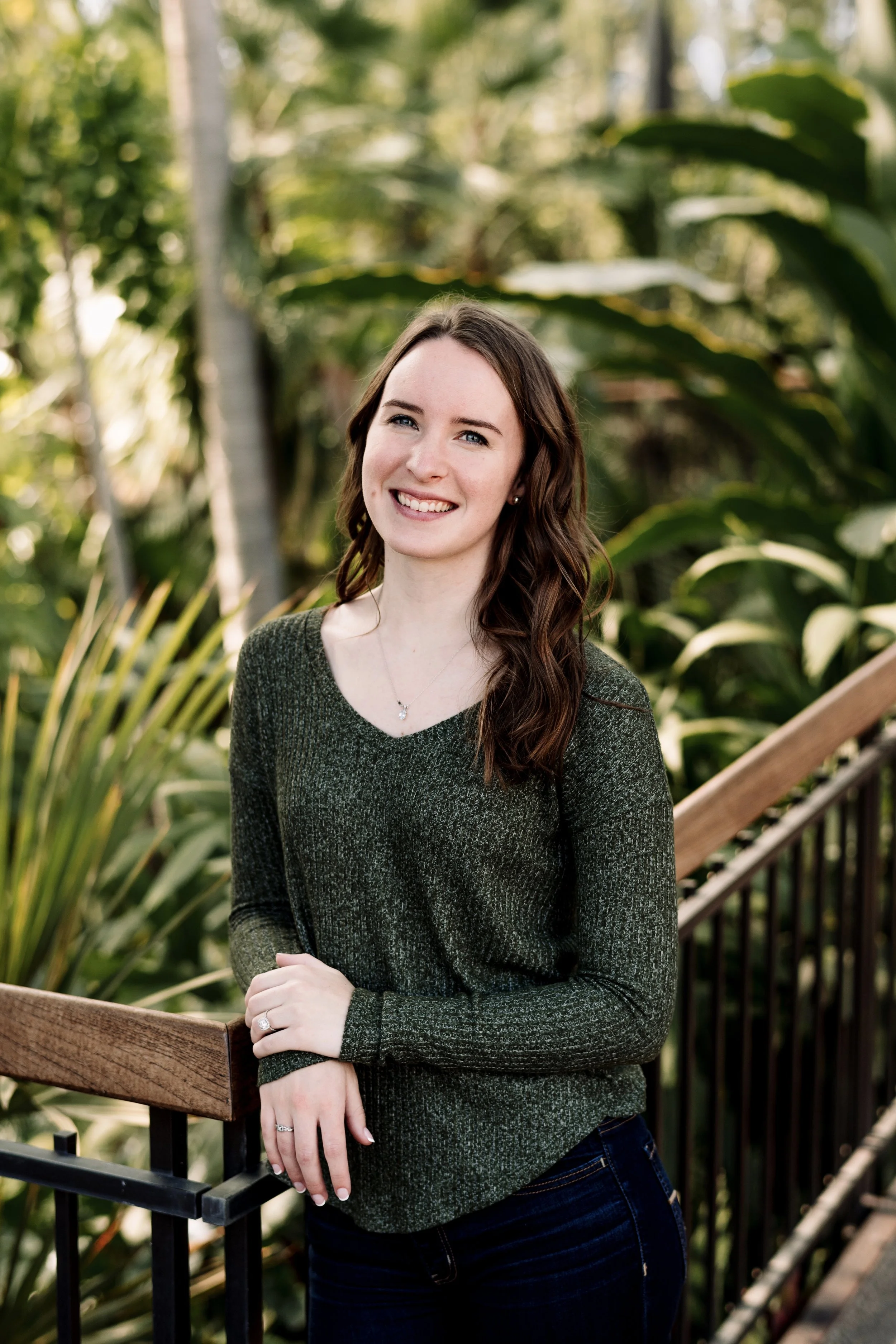 A young woman with long brown hair, wearing a dark green sweater and dark jeans, standing outdoors near a wooden railing, smiling at the camera with green foliage in the background.