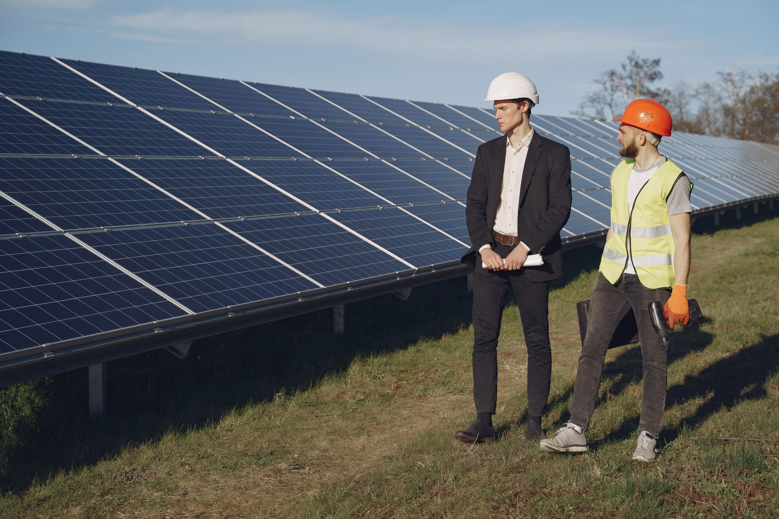 Two men wearing safety helmets and work vests inspecting solar panels in a field.