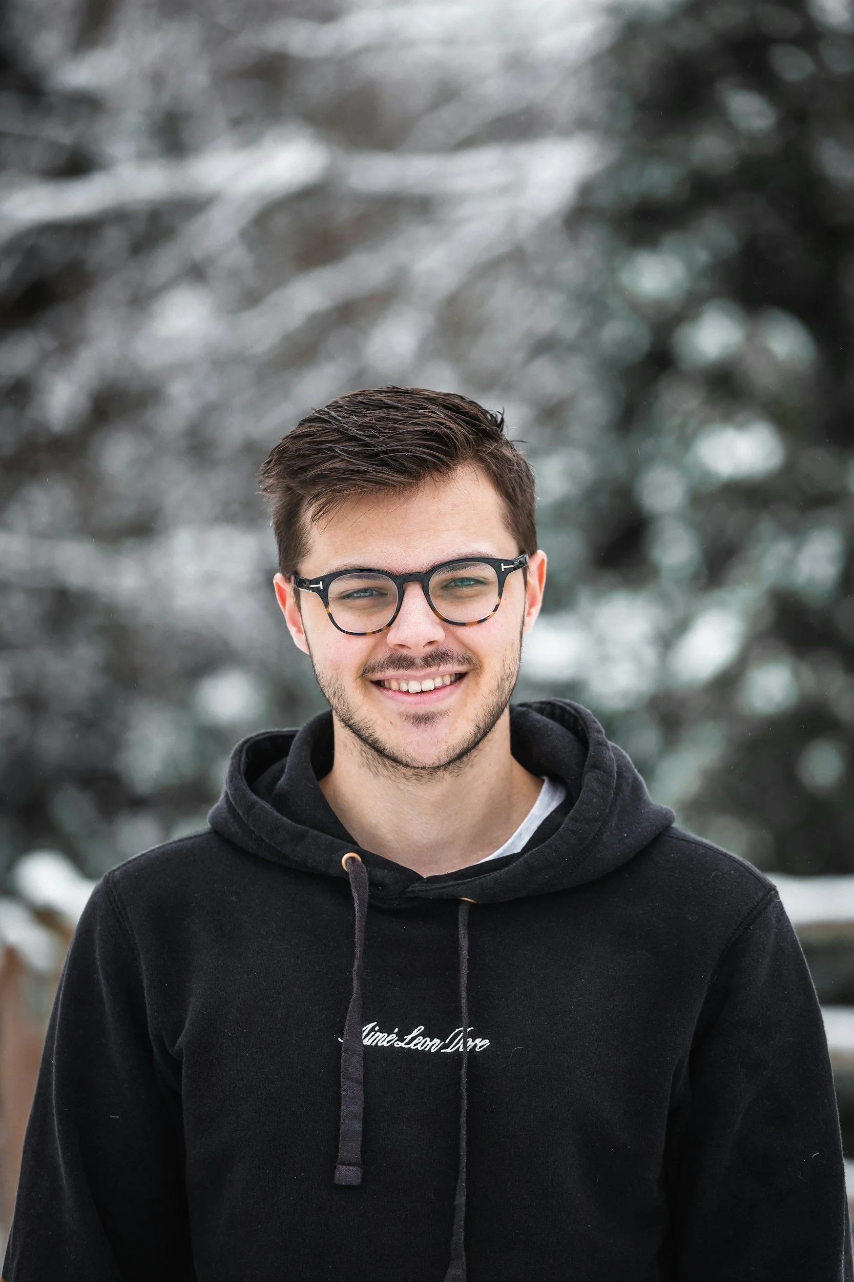 A young man with short brown hair, glasses, and a beard is smiling, standing outdoors in front of snow-covered trees.