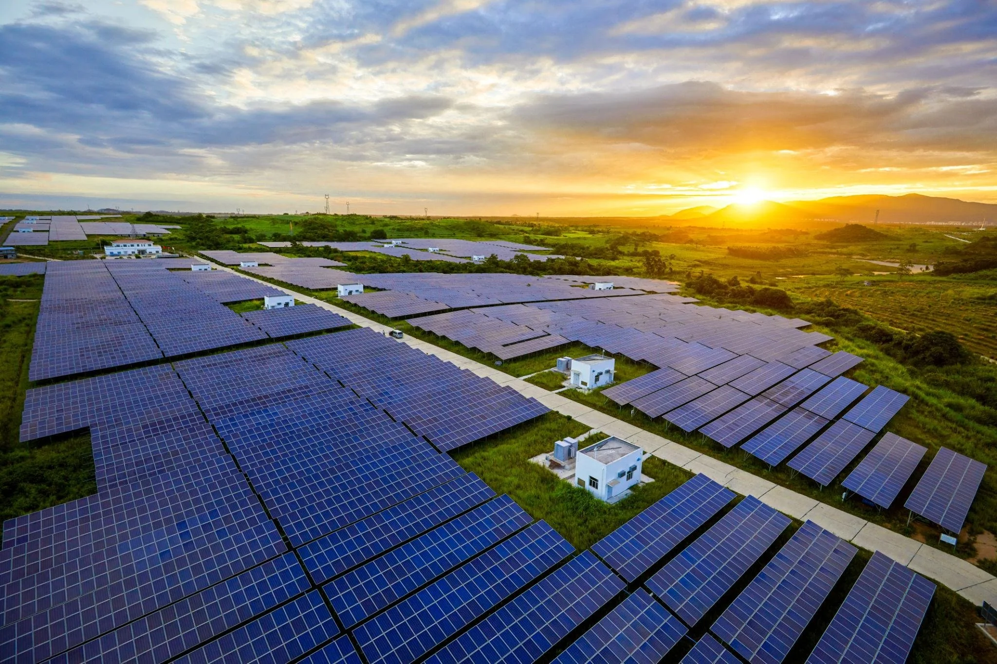 An expansive solar farm with rows of solar panels on green fields under a sunset sky with clouds.