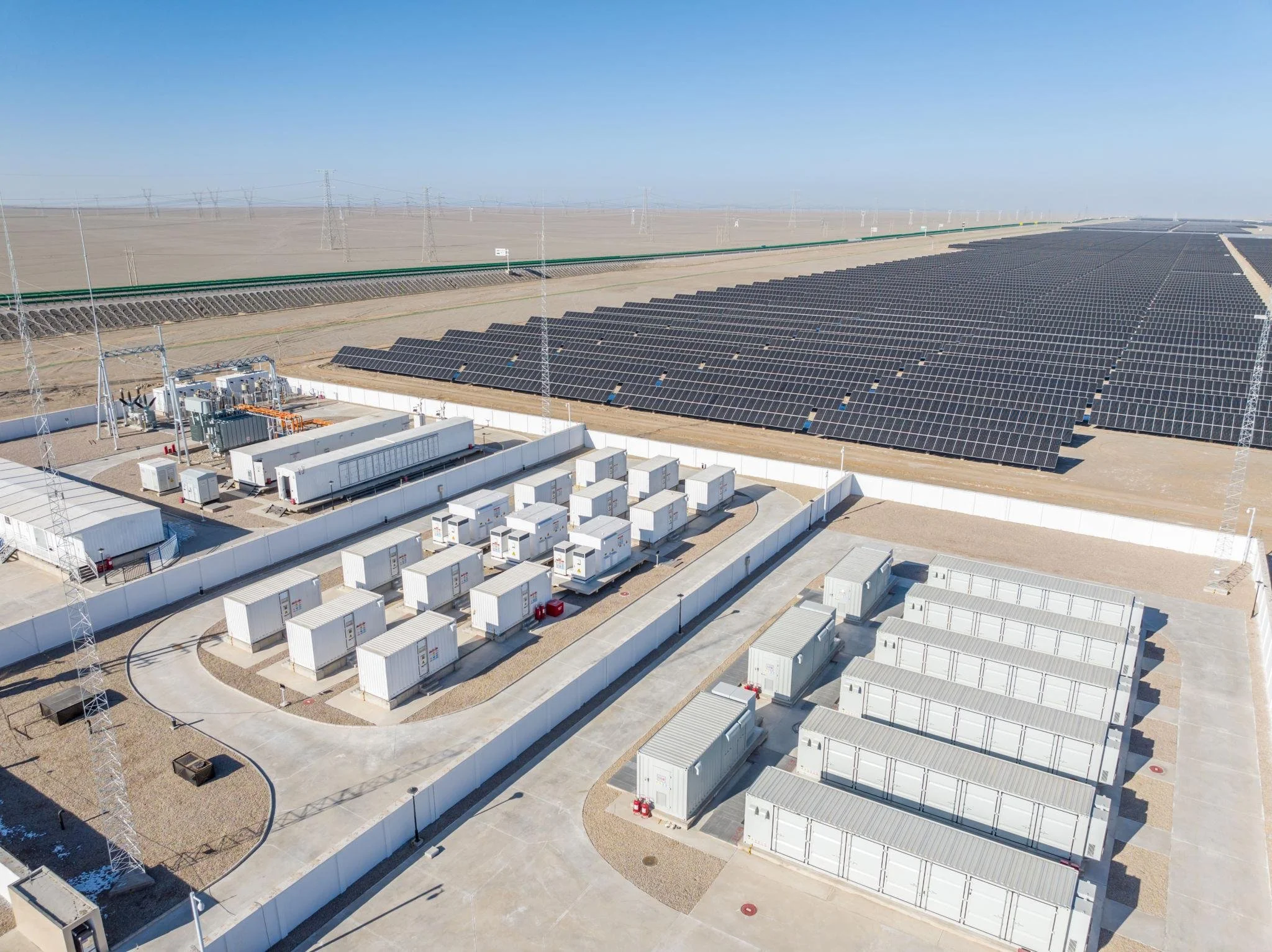 A large solar power farm with rows of solar panels installed on the ground, surrounded by white fencing. There are electrical equipment and control buildings nearby, with a clear blue sky overhead.