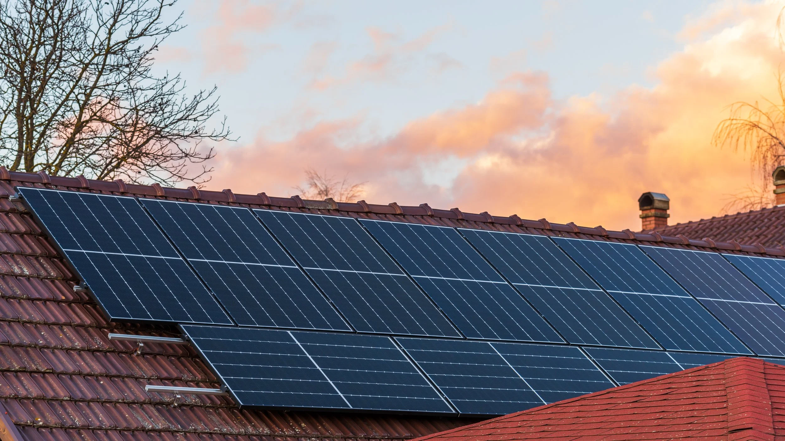 Solar panels installed on a red tiled roof during sunset with trees and a partly cloudy sky in the background.