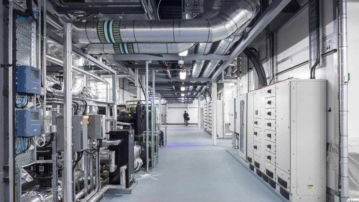 Interior of an industrial facility with machinery, control panels, and ductwork, including a person walking in the background.