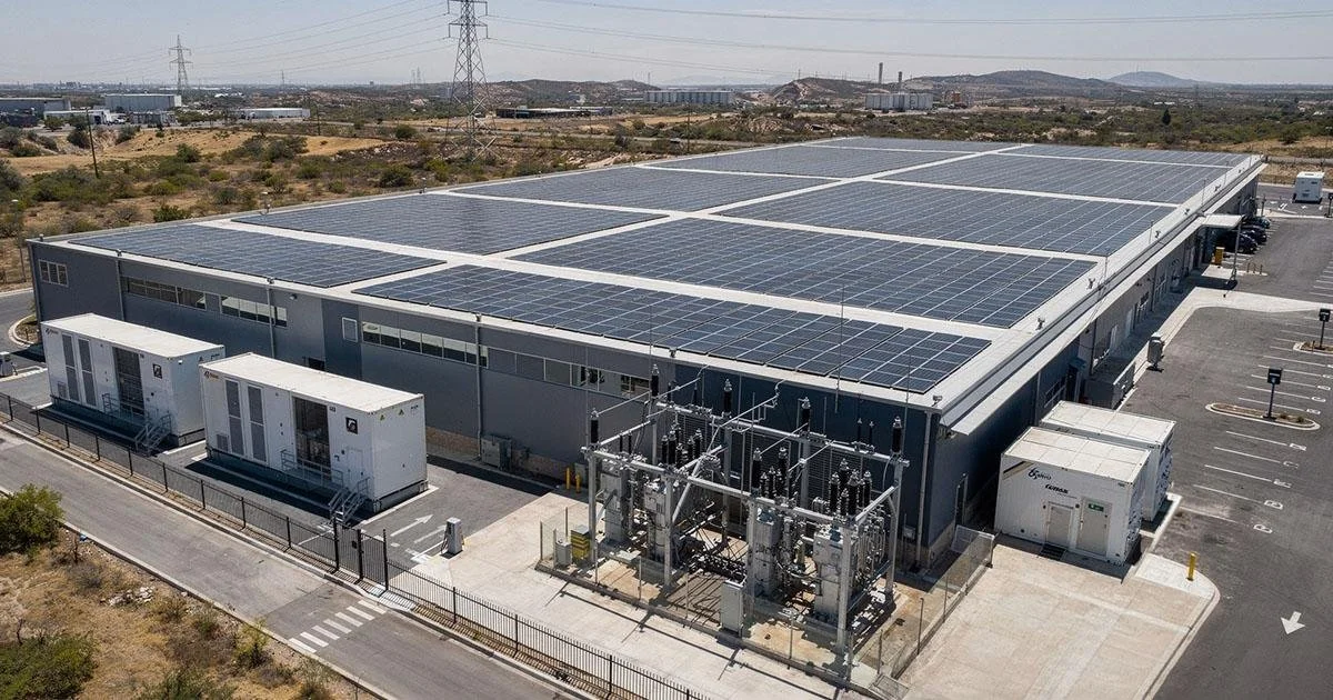 Large industrial building with a roof covered in solar panels, surrounded by parking lots and electrical infrastructure in a semi-arid landscape.