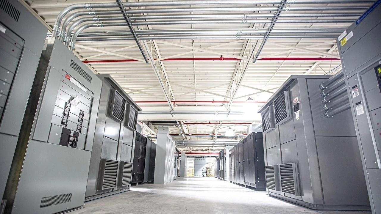 Interior of a data center with server racks and electrical equipment arranged in rows under a white ceiling with exposed pipes and conduits.