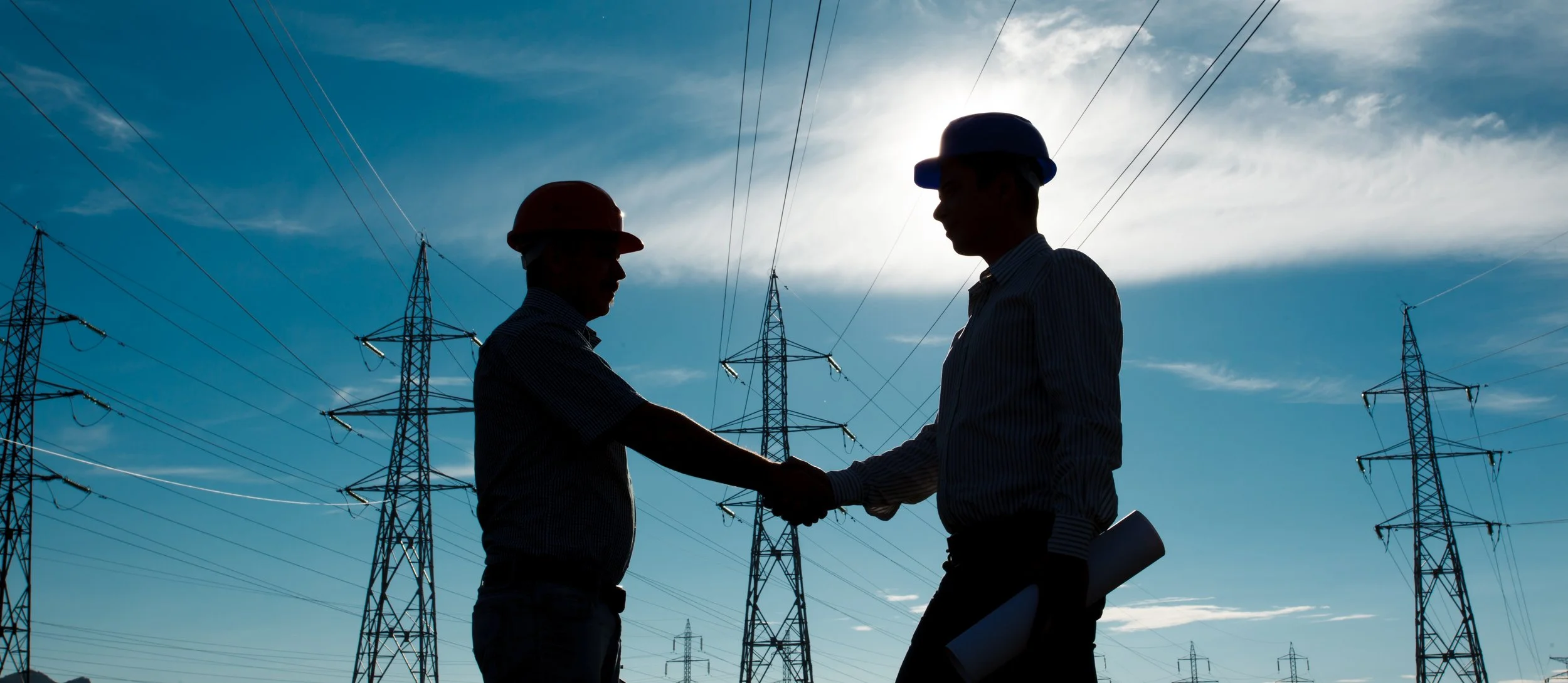 Two engineers in safety helmets shaking hands under high-voltage power lines with pylons against the sky.