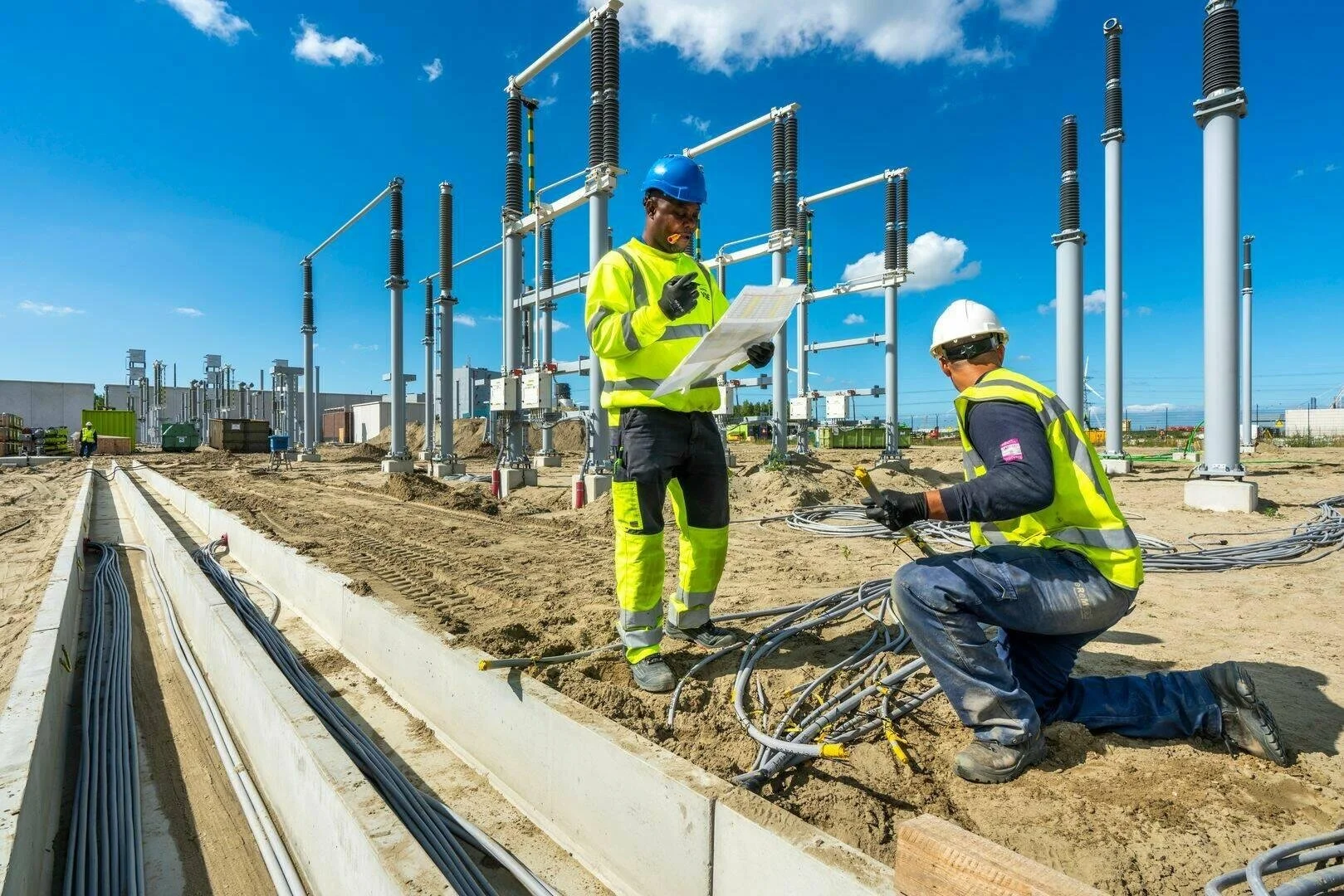 Two construction workers in high-visibility clothing and hard hats working on electrical wiring at an outdoor construction site under blue sky.