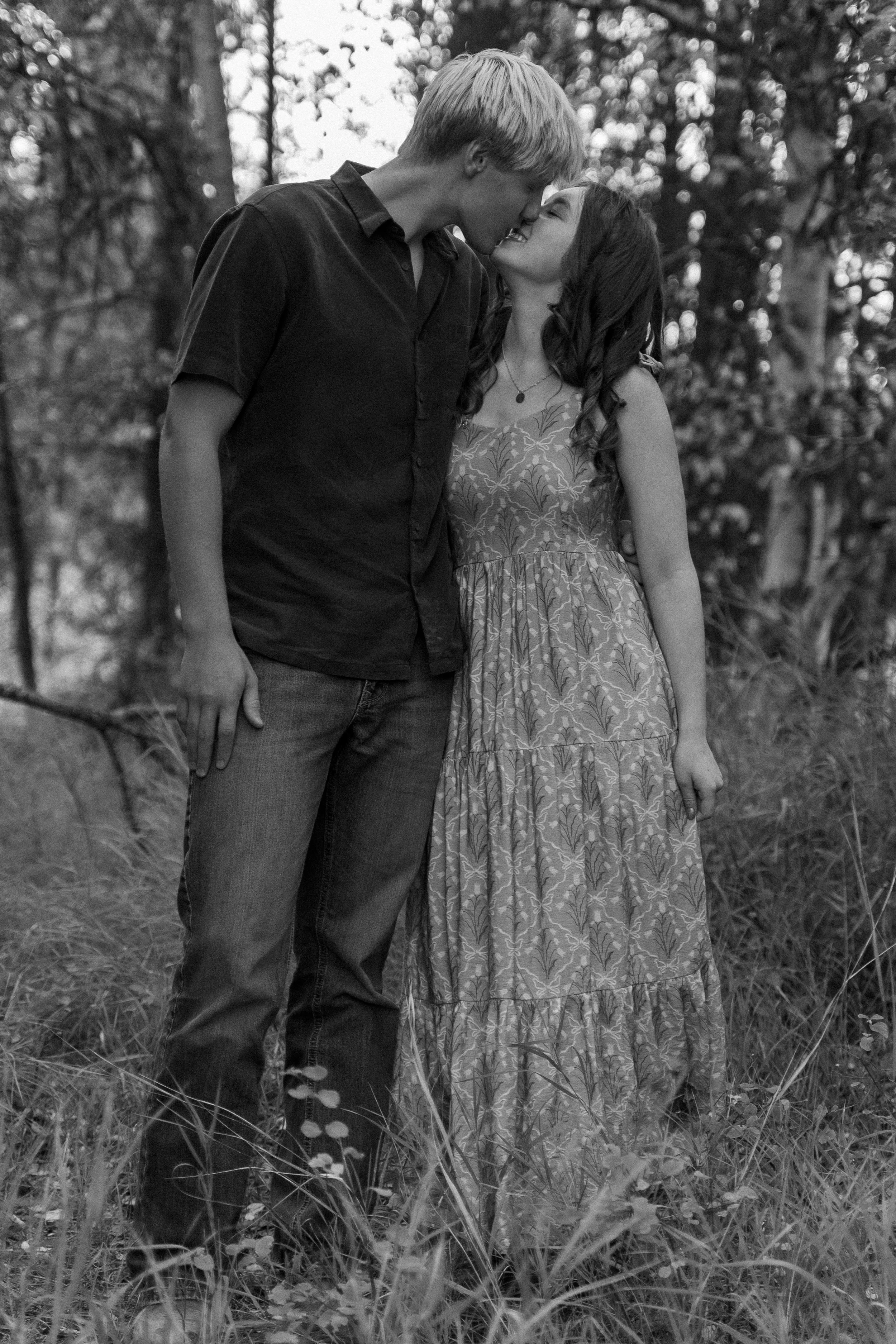 Black-and-white portrait of a smiling couple embracing and kissing in a sunlit forest.