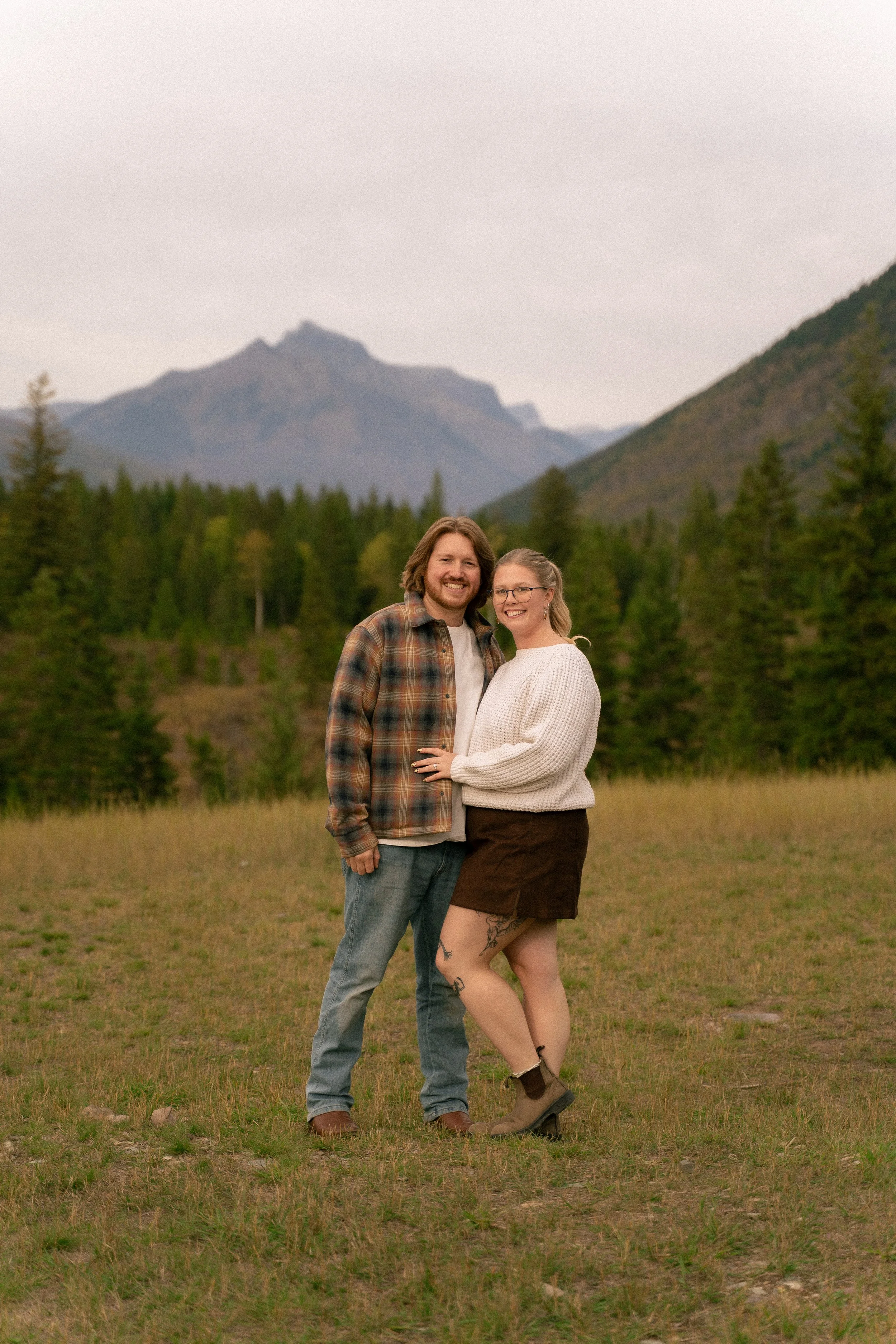 Smiling couple embracing in a grassy field with a forest and rugged mountain peak in the background