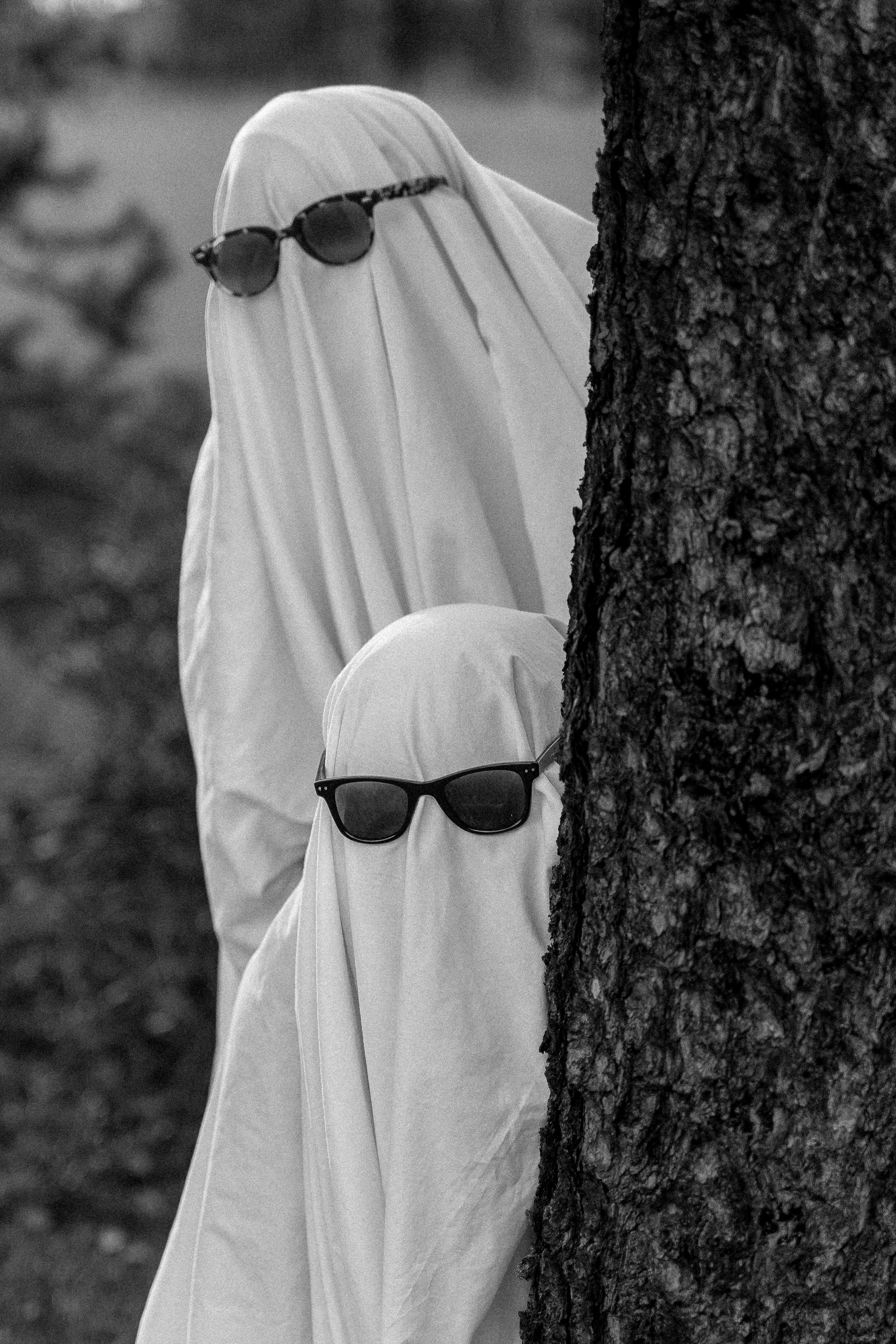 Black-and-white photo of two people in ghost costumes wearing sunglasses and peeking from behind a tree