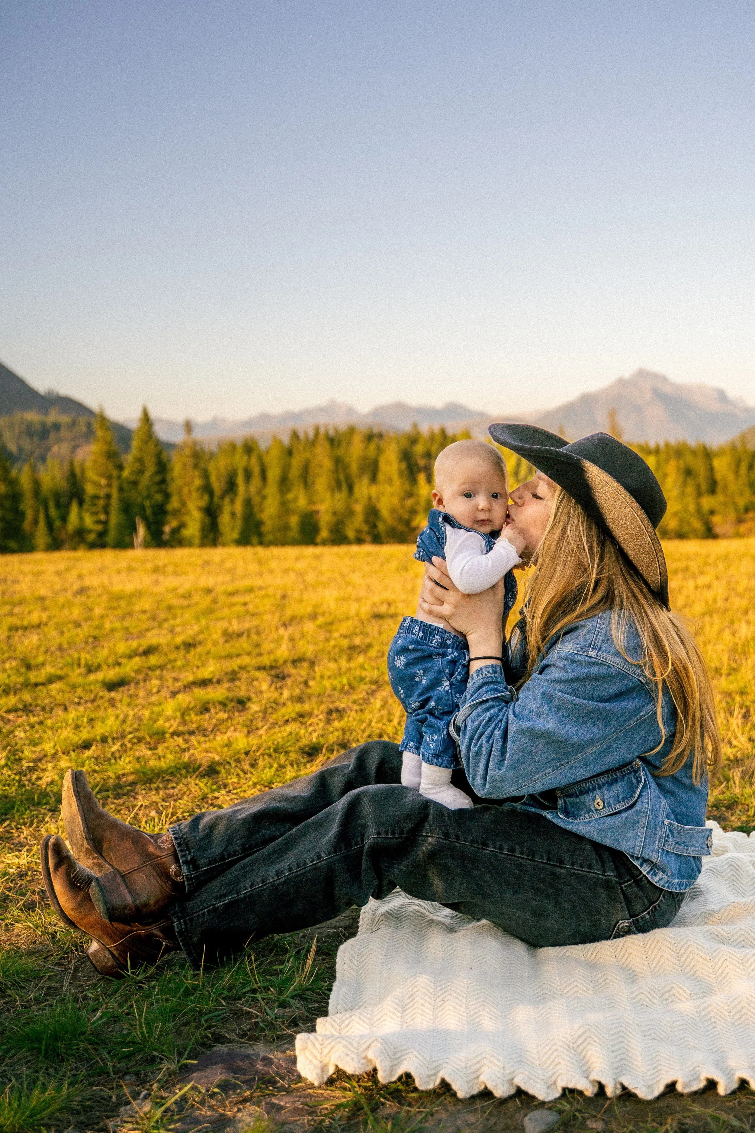 Mother kissing her baby in a sunlit mountain meadow, surrounded by autumn trees and mountains.