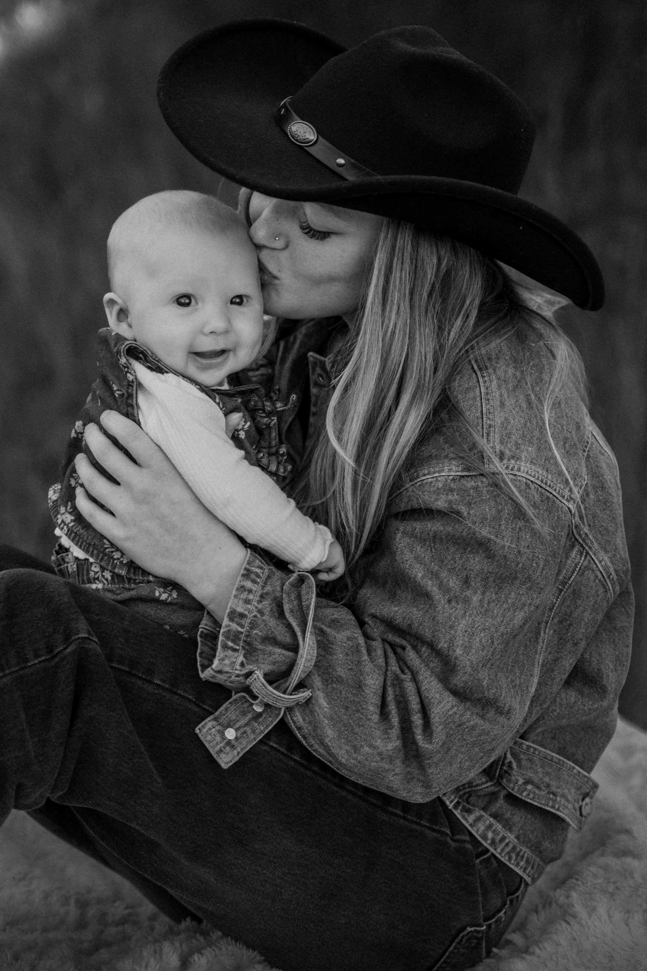 Black-and-white portrait of a smiling baby in a floral sling as a woman in a wide-brimmed hat kisses the baby's cheek.