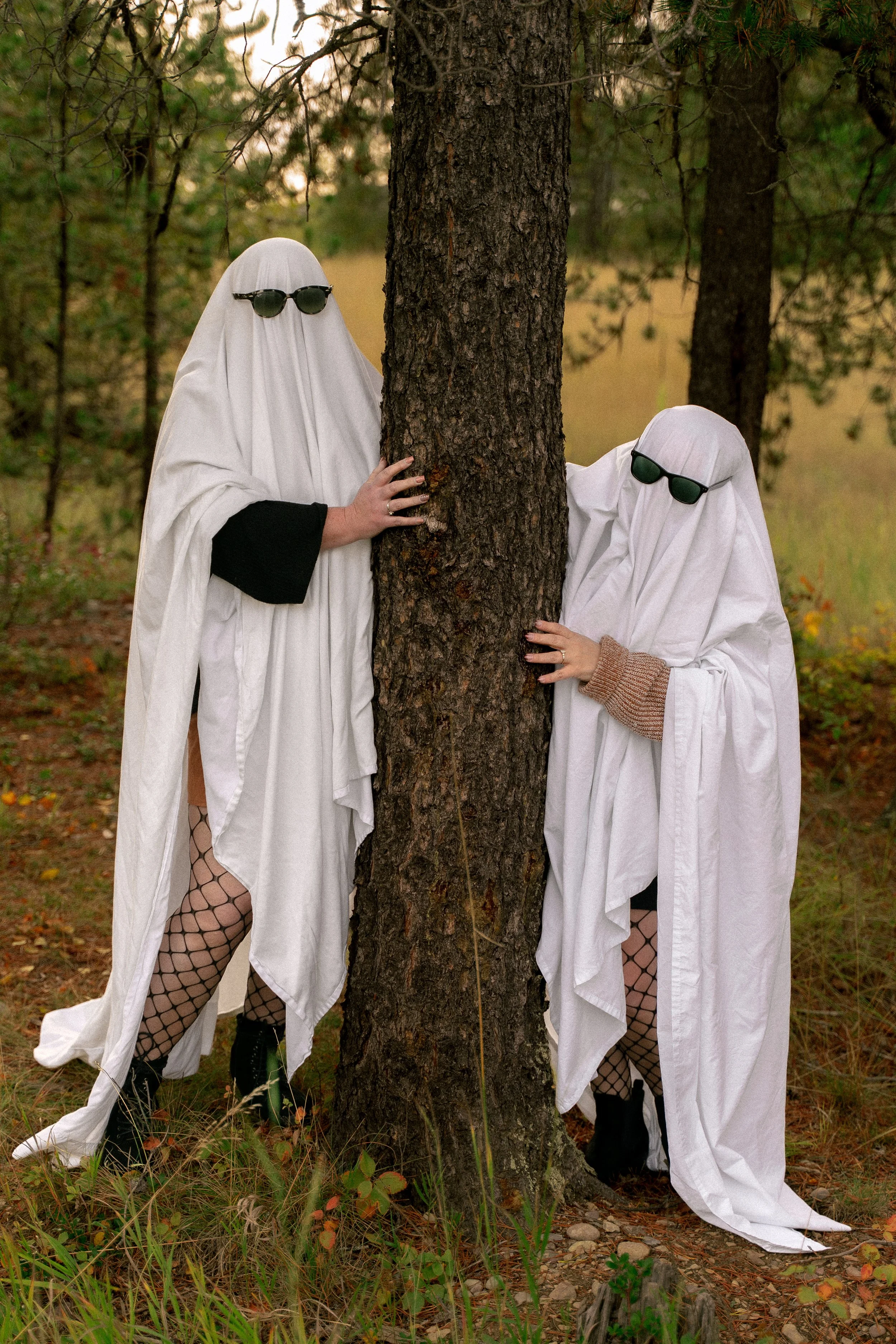 Two people dressed as ghosts and wearing sunglasses pose around a pine tree in a forest.