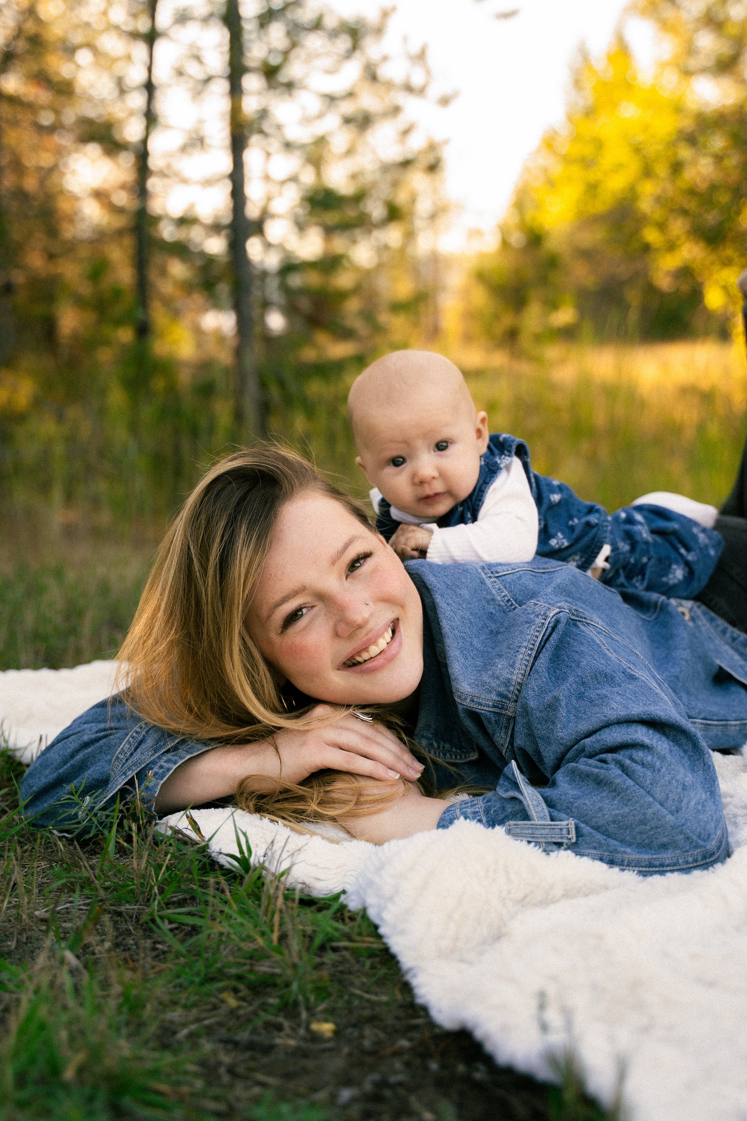 Smiling mother in a denim jacket holding her baby outdoors on a blanket among autumn trees