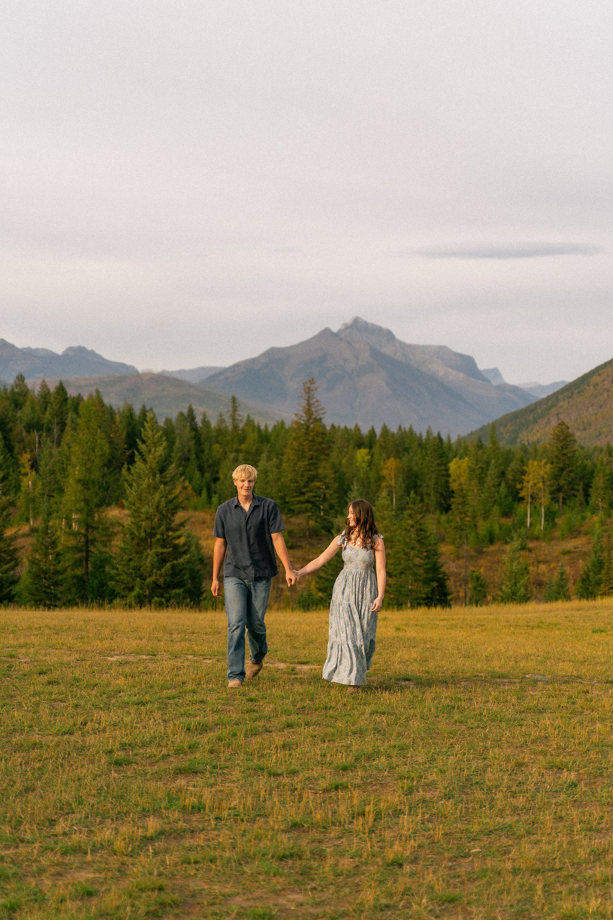 Couple holding hands walking through a meadow with evergreen forest and rugged mountains in the background