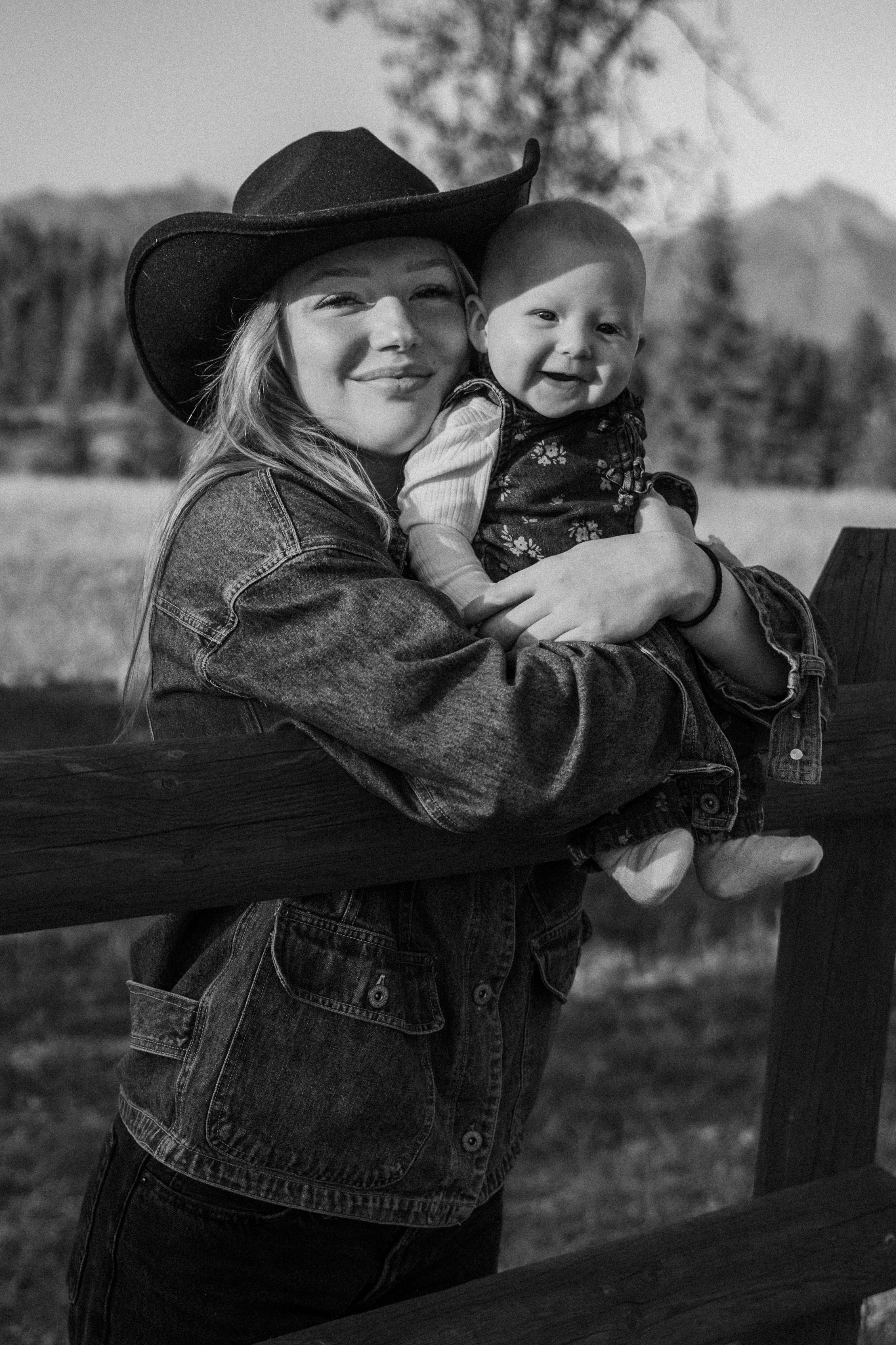 Black-and-white portrait of a smiling woman in a cowboy hat holding a baby outdoors.