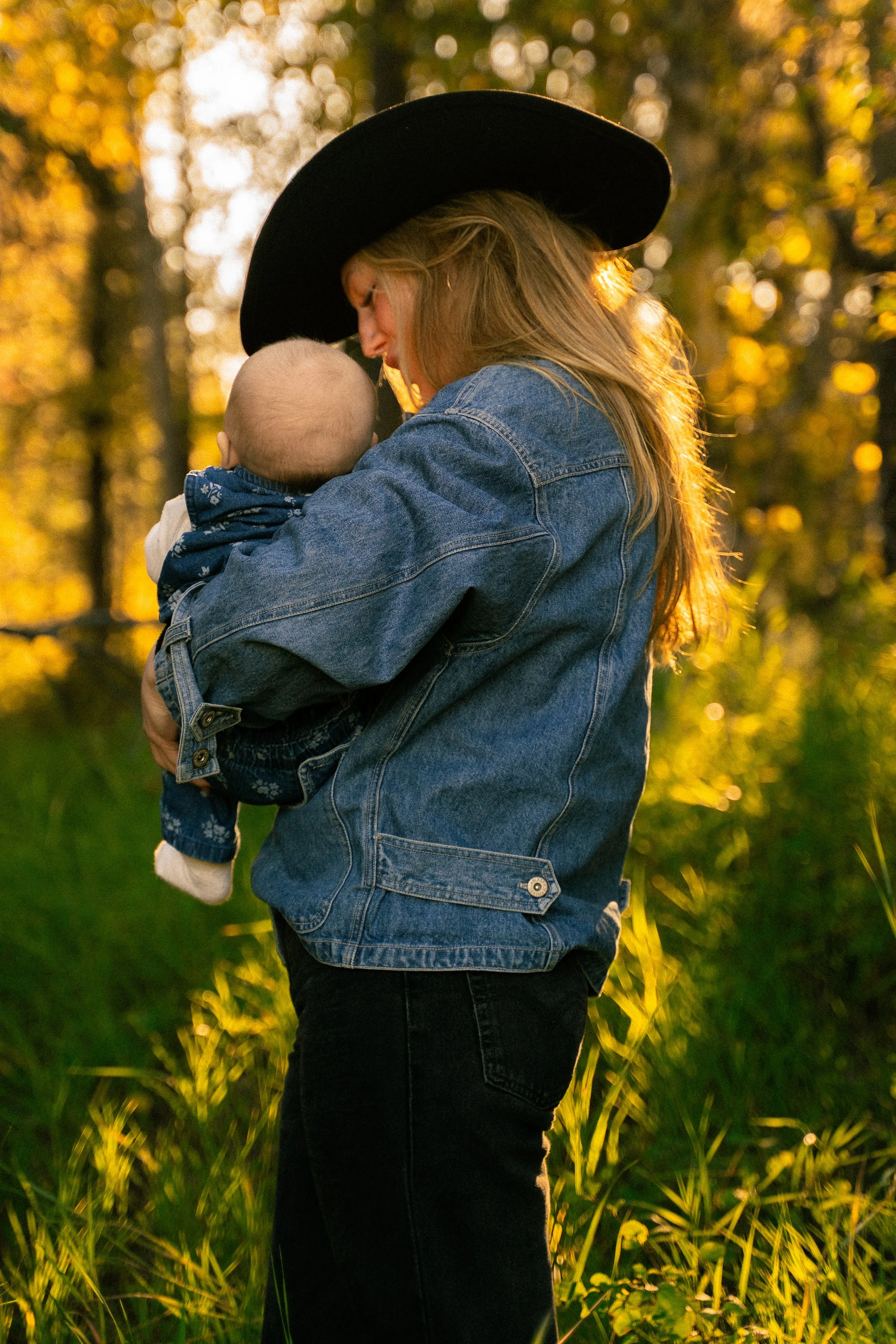 Mother in a black hat and denim jacket holding a baby outdoors in warm sunlight