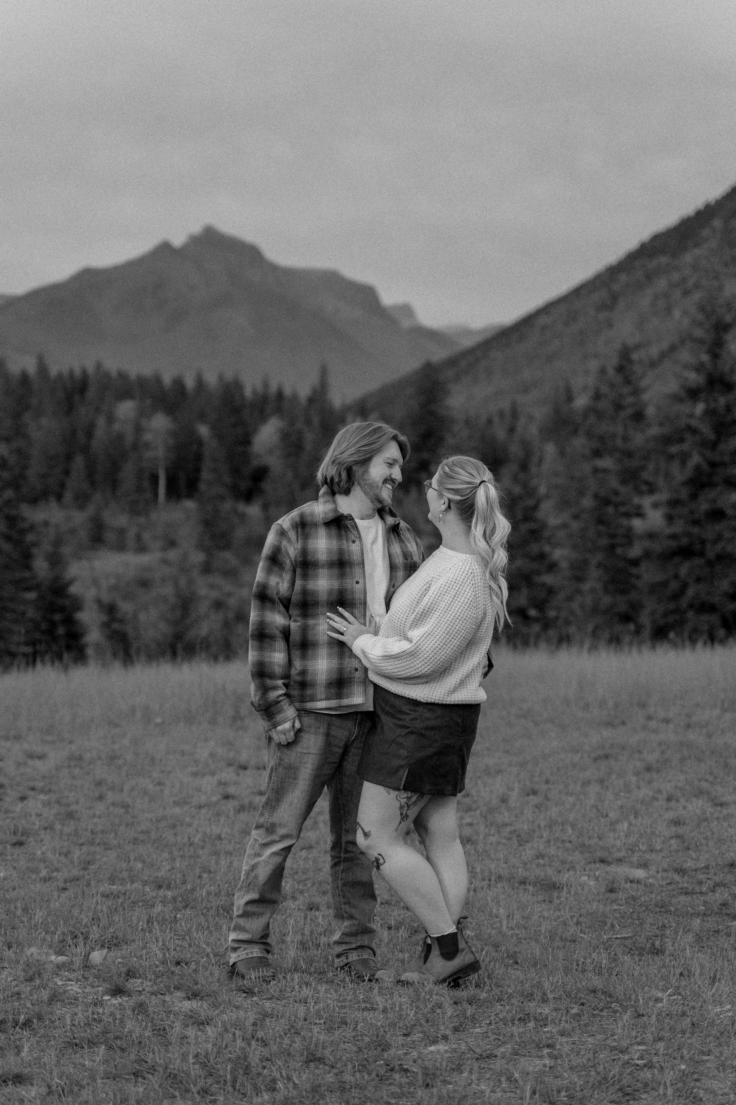 Black-and-white photo of a smiling couple embracing in a grassy field with mountain peaks and forest behind them