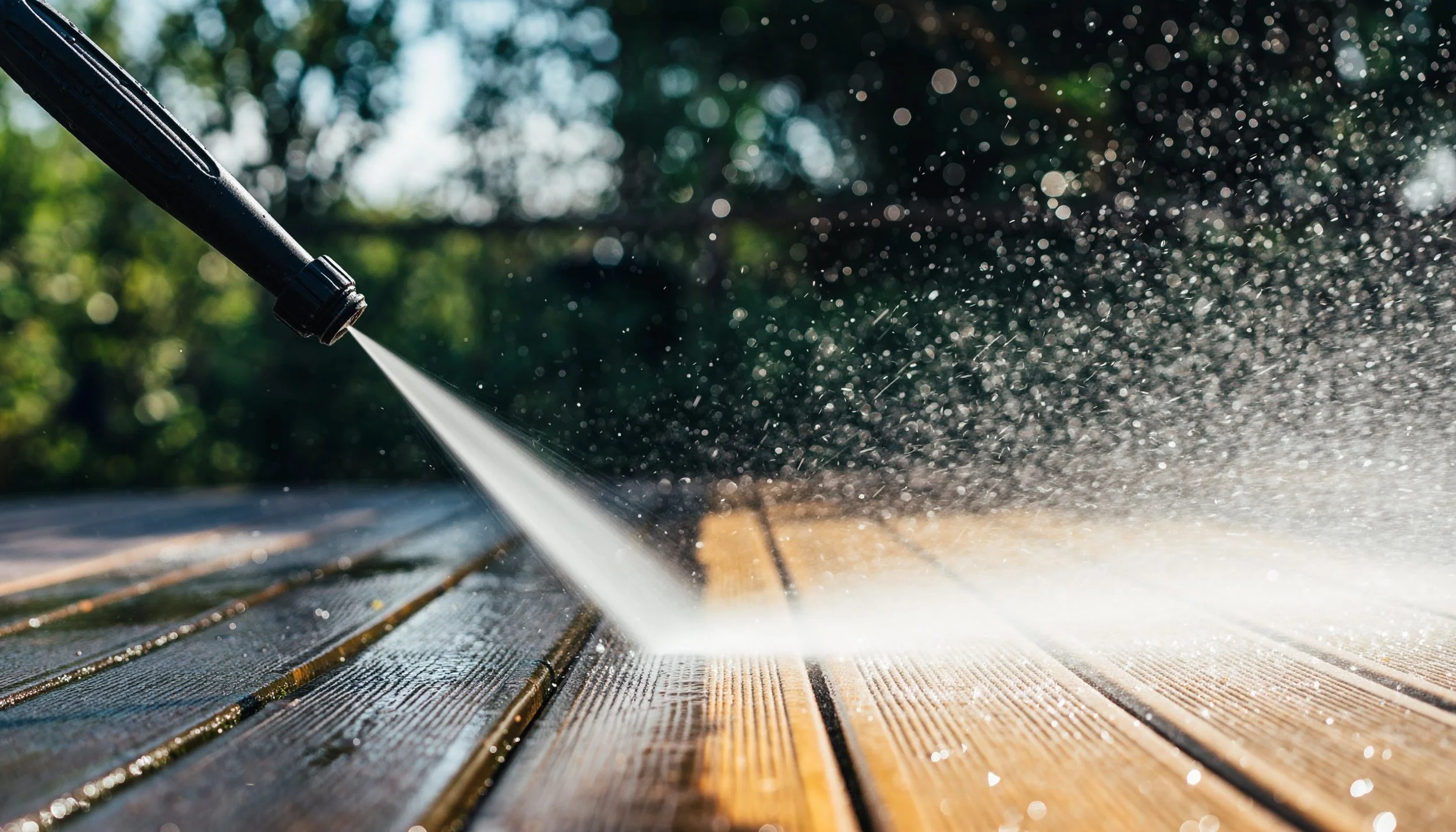 Pressure washing a wooden deck with sunlight filtering through trees in the background.