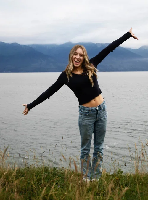 A young woman standing outdoors by a body of water, with mountains in the background, smiling and spreading her arms wide.