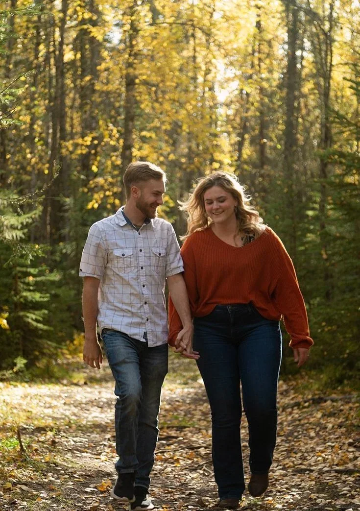 A young couple holding hands and walking through a forest trail during autumn, with golden leaves and sunlight filtering through the trees.